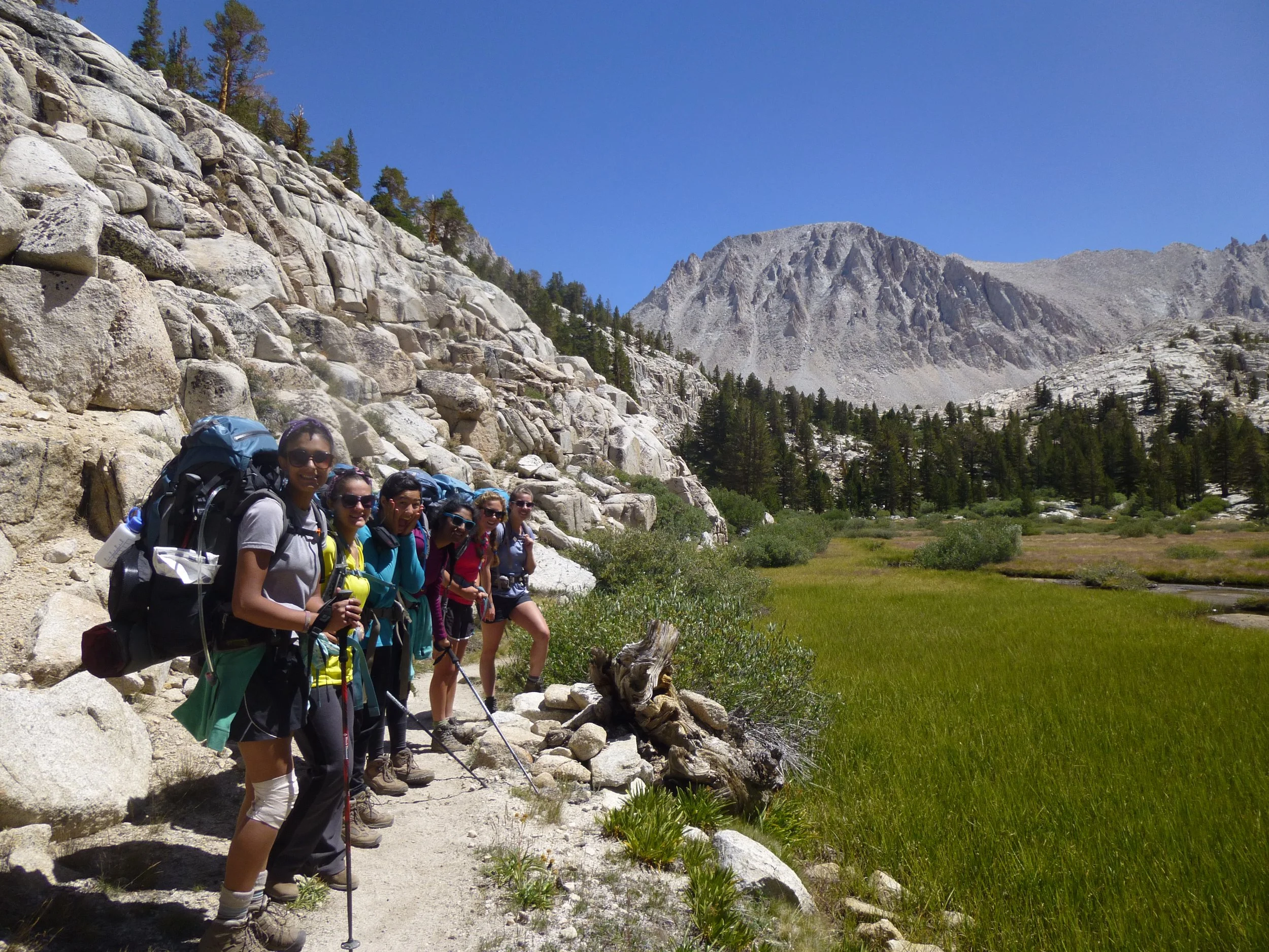 Girl Scout backpackers on a trail with a rock wall to their left, a meadow to their right, and pine trees, mountains, and a blue sky in the distance.