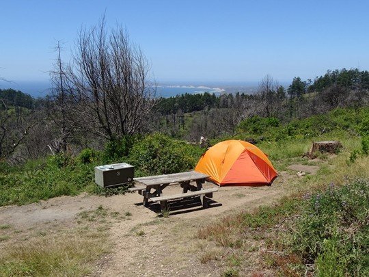 An orange tent set-up at a campsite on a ridge with the pacific ocean in the background.