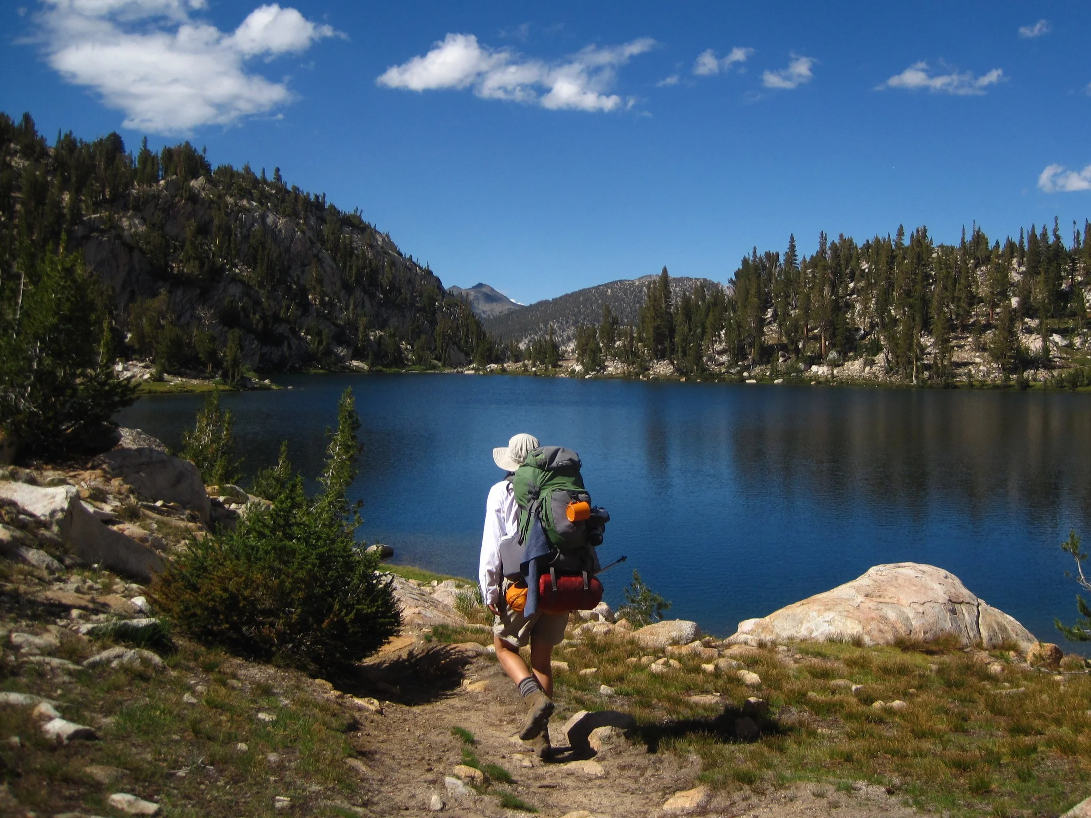 A Girl Scout backpacker hiking on the edge of a turquoise lake with rolling hills covered with pine trees in the distance.
