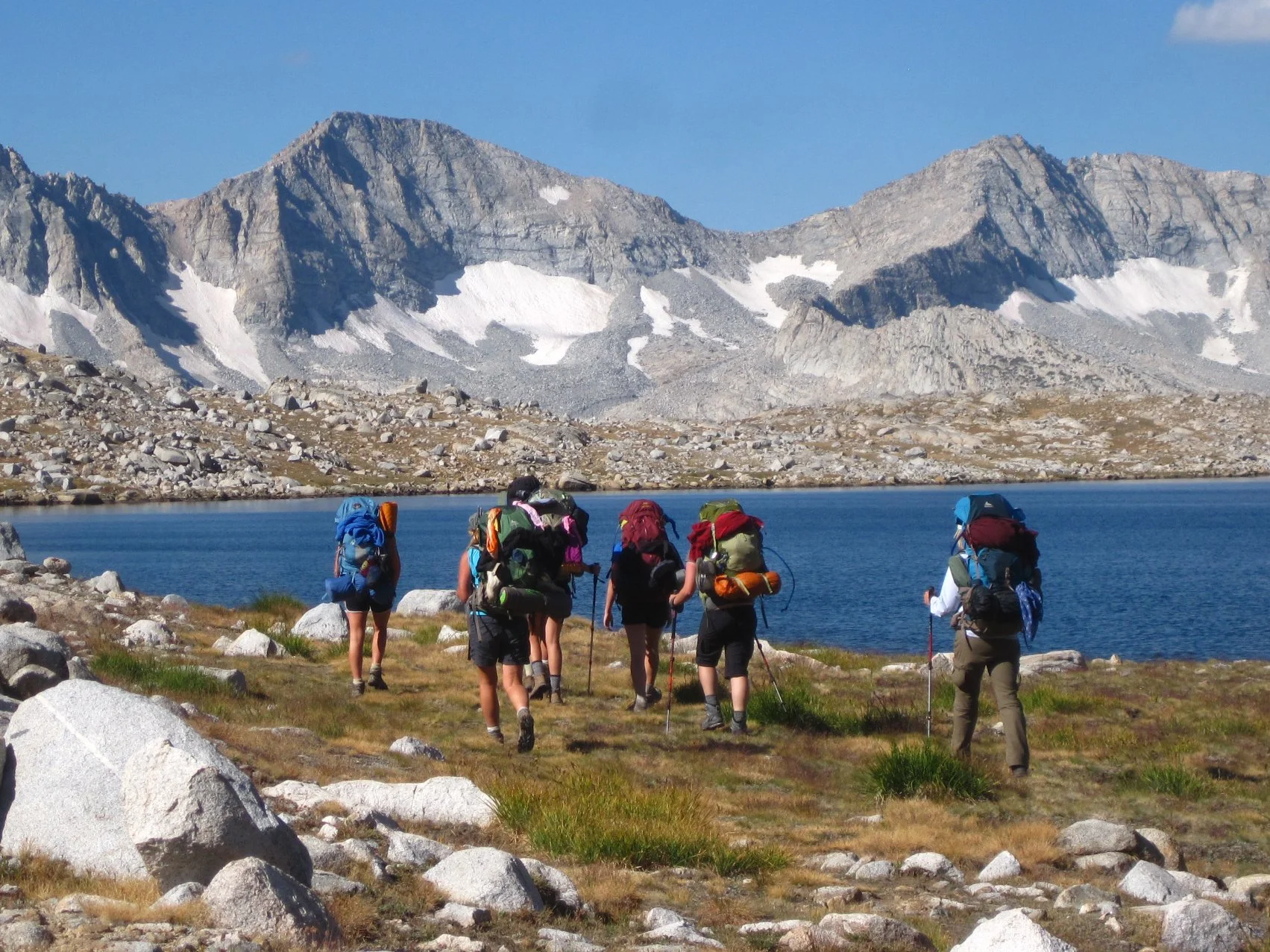 A group of 8 Girl Scout backpackers hiking on the edge of an alpine lake with snow dotted mountains and a blue sky in the background.