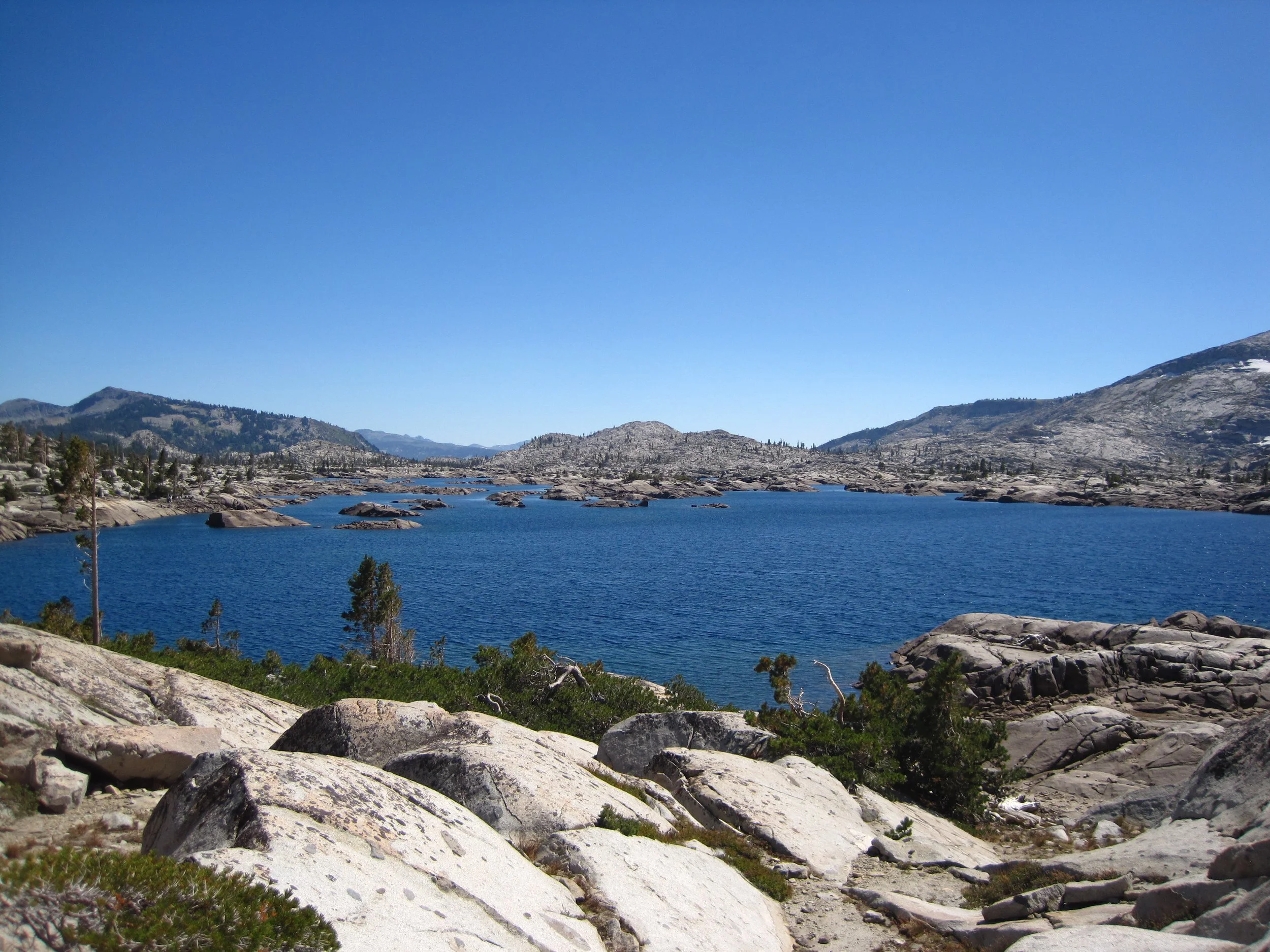 A blue lake with white rocks and green shrubbery in the foreground with mountains in the background.