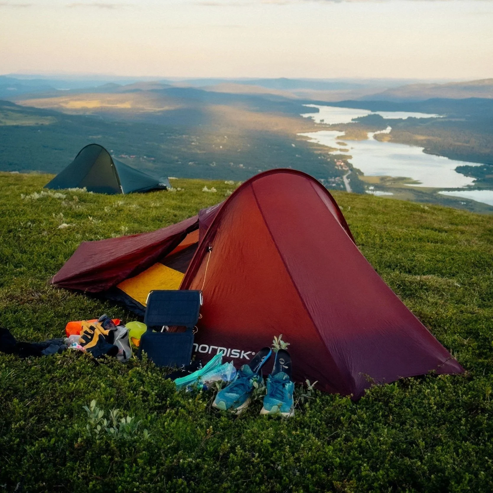 A red tent on green grass with hiking shoes, and other equipment strewn about; a blue tent is in the background as well as a canyon river deep in the background.