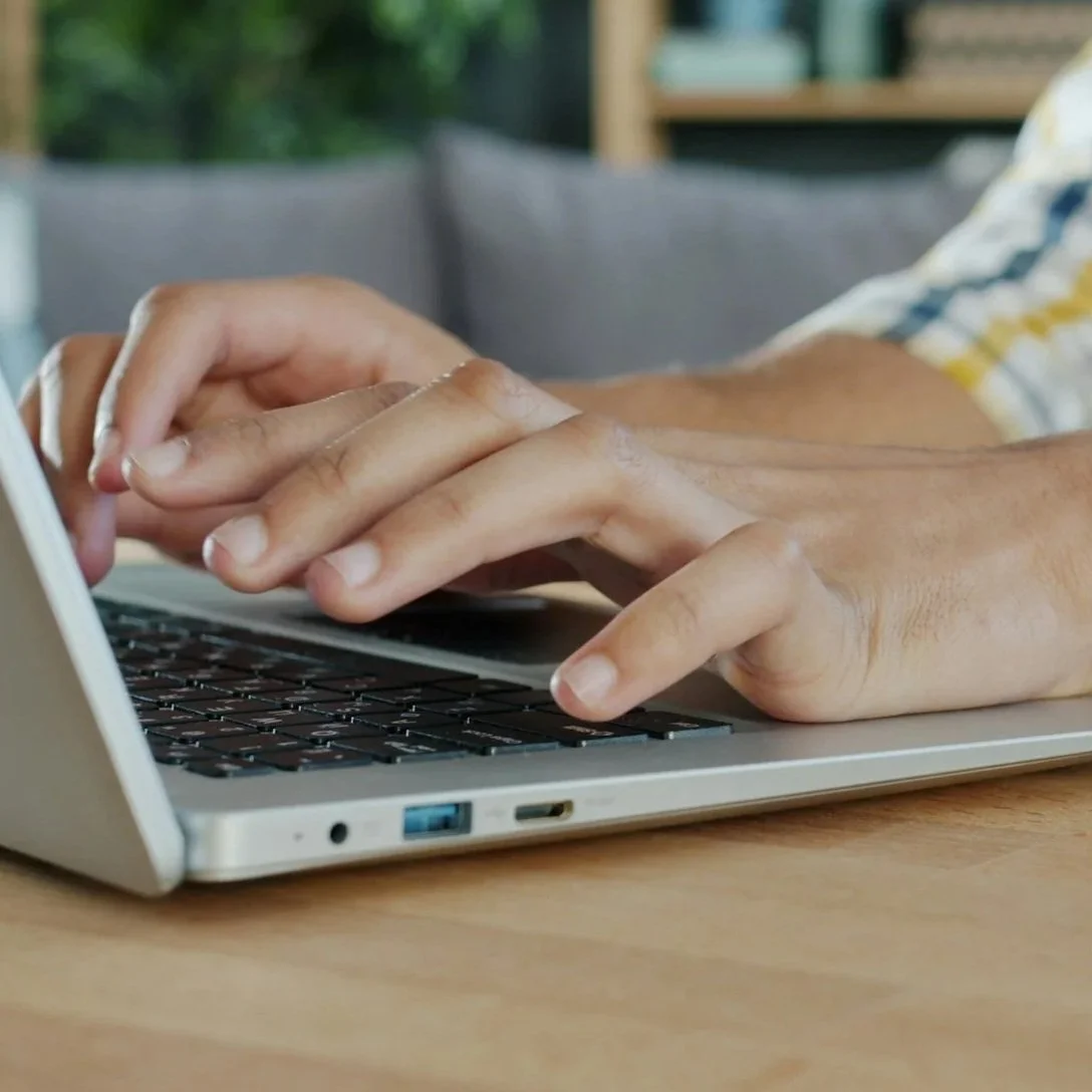 Hands of a person offscreen typing on a laptop keyboard on a pale, wooden surface.