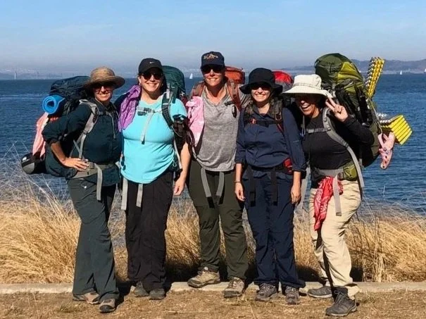 Five adult Girl Scouts with backpacks on standing on the water's edge with dry brush waving in the wind.