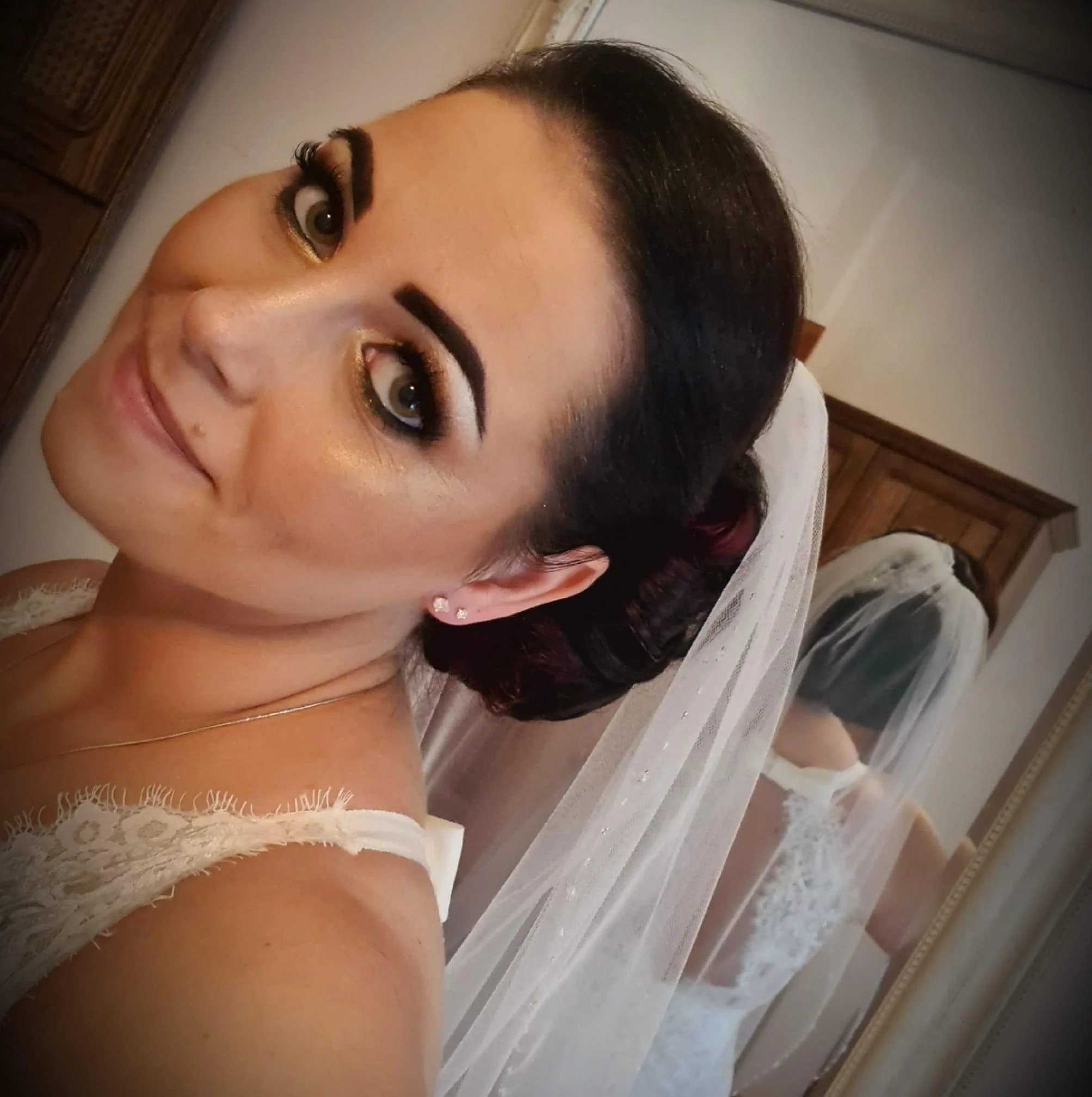 A woman with dark hair styled in an updo, wearing makeup, earrings, and a white lace dress with a veil, taking a selfie in front of a mirror.