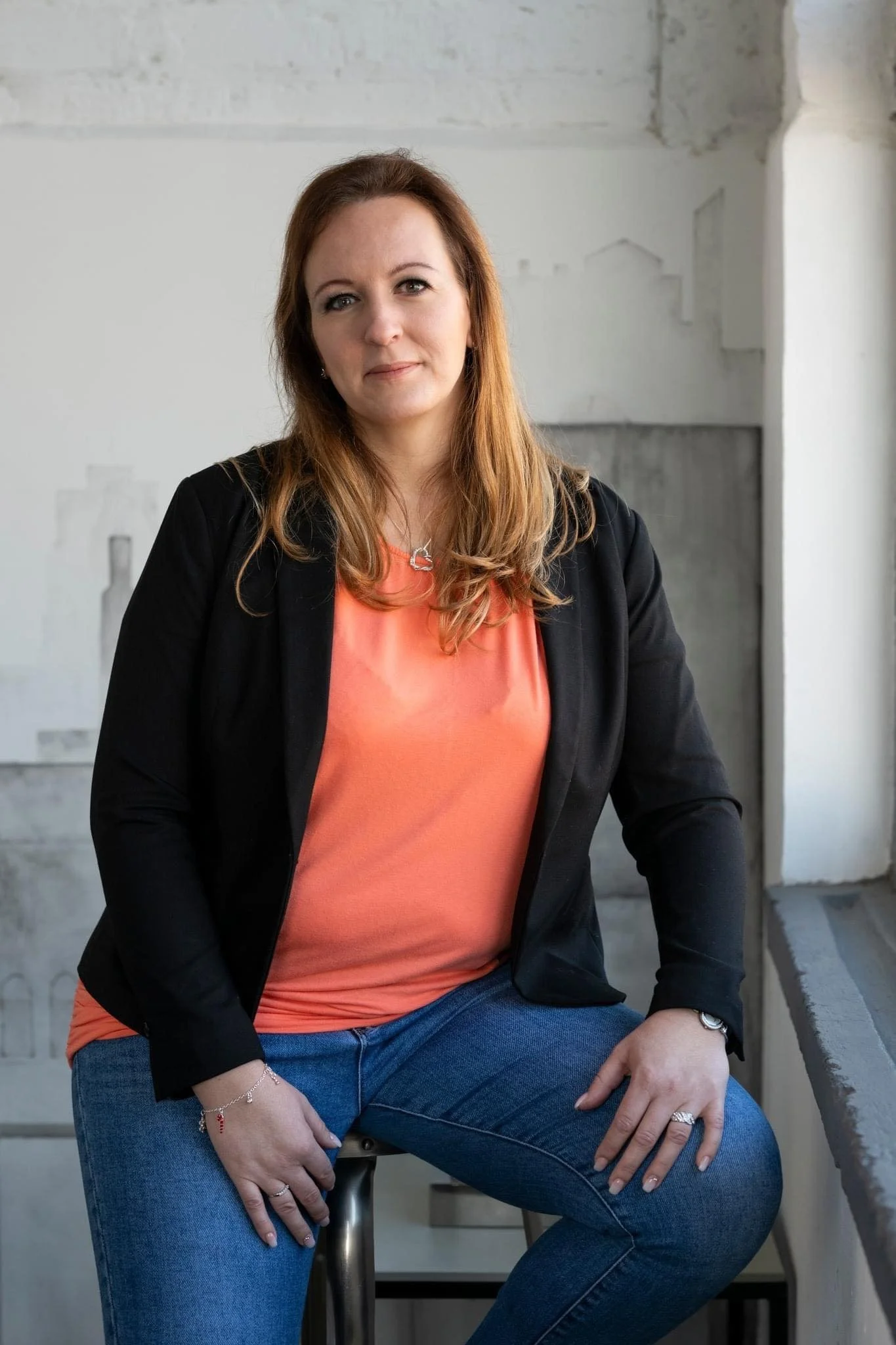 A woman with long brown hair wearing a black blazer, coral top, and jeans, sitting on a stool in a modern, minimalist space with concrete and white walls.