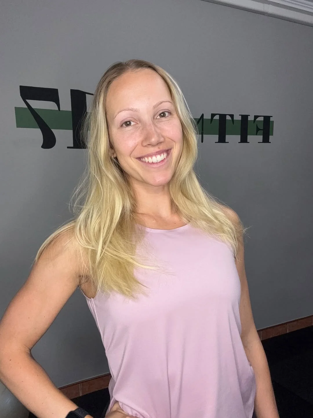 A smiling woman with long blonde hair wearing a pink sleeveless top standing in front of a gray wall with the letters 'FIT' partially visible.
