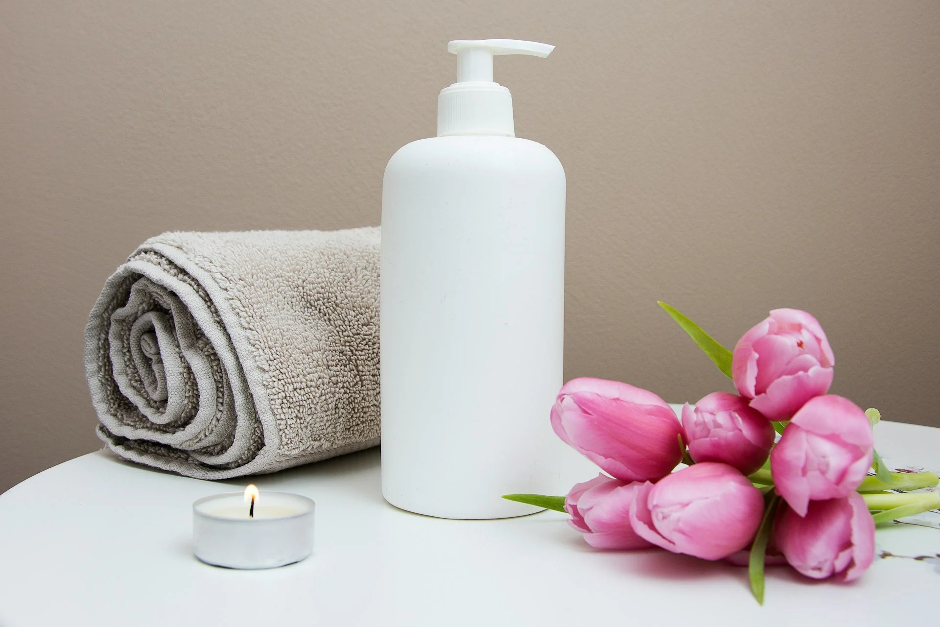 Rolled beige towel, white lotion pump bottle, small lit tealight candle, pink tulips with green leaves on a white surface against a beige wall.
