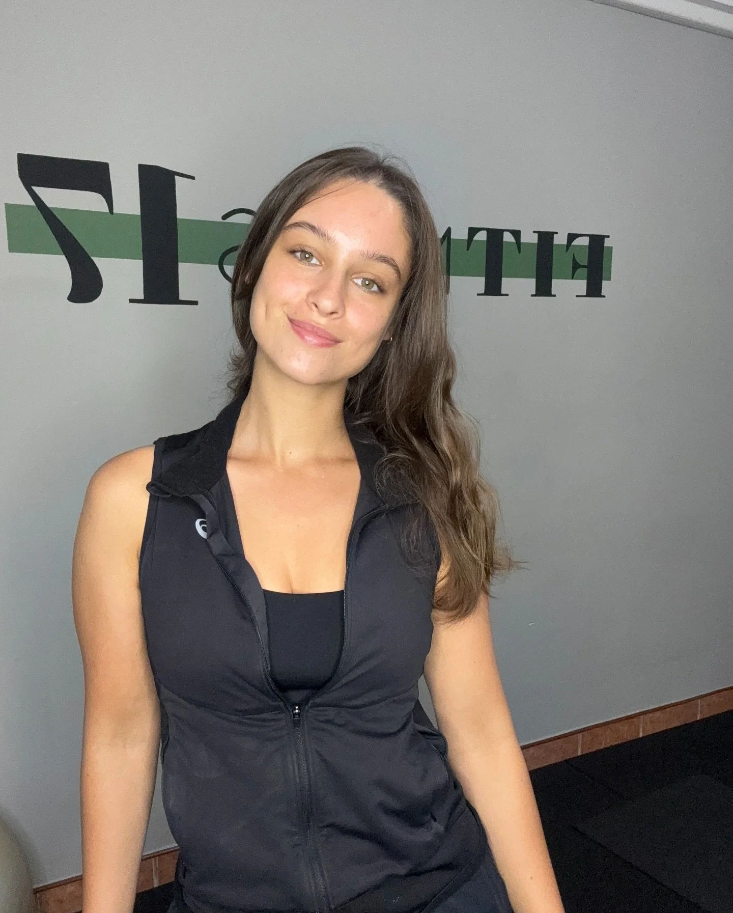 A young woman with long brown hair, wearing a black athletic vest and black tank top, smiling in front of a light gray wall with partially visible black and green lettering.