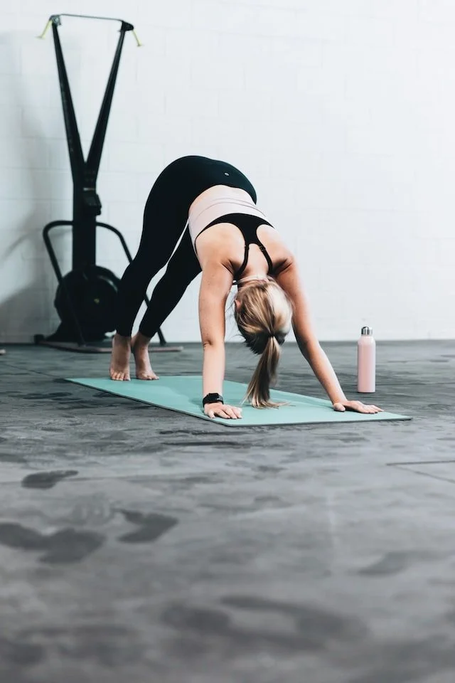 A woman practicing yoga in a gym, in downward dog pose, with a water bottle nearby.