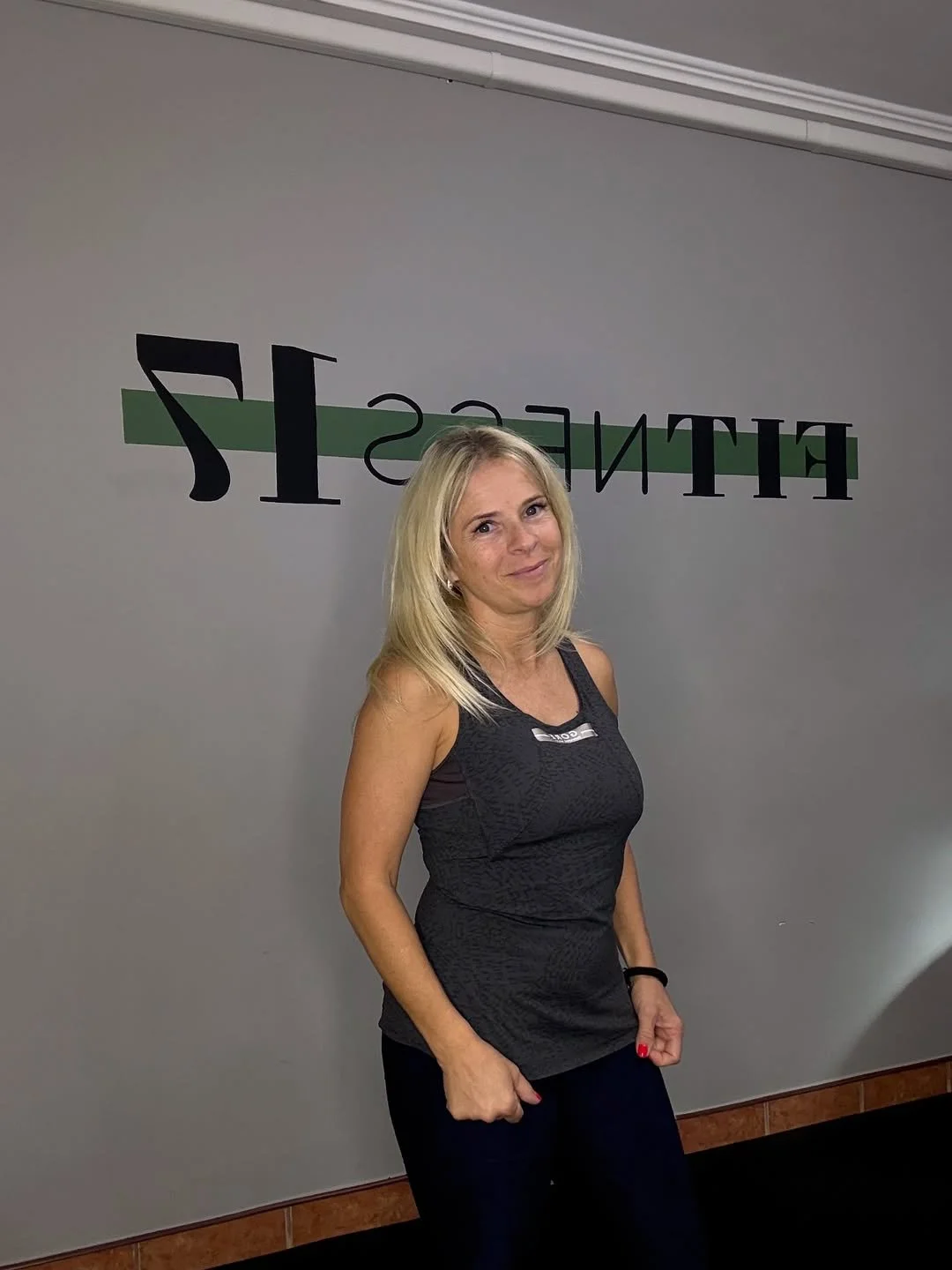 Woman with blonde hair standing in front of a wall with the word 'FITNESS' mirrored, wearing a black sleeveless athletic top and black pants, smiling.