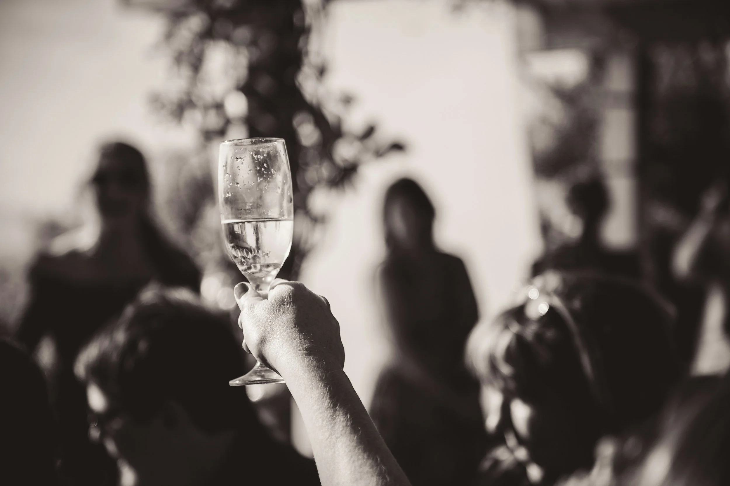 A person holding a champagne flute with bubbles in it, raised in a toast, with blurred people in the background at a social gathering.