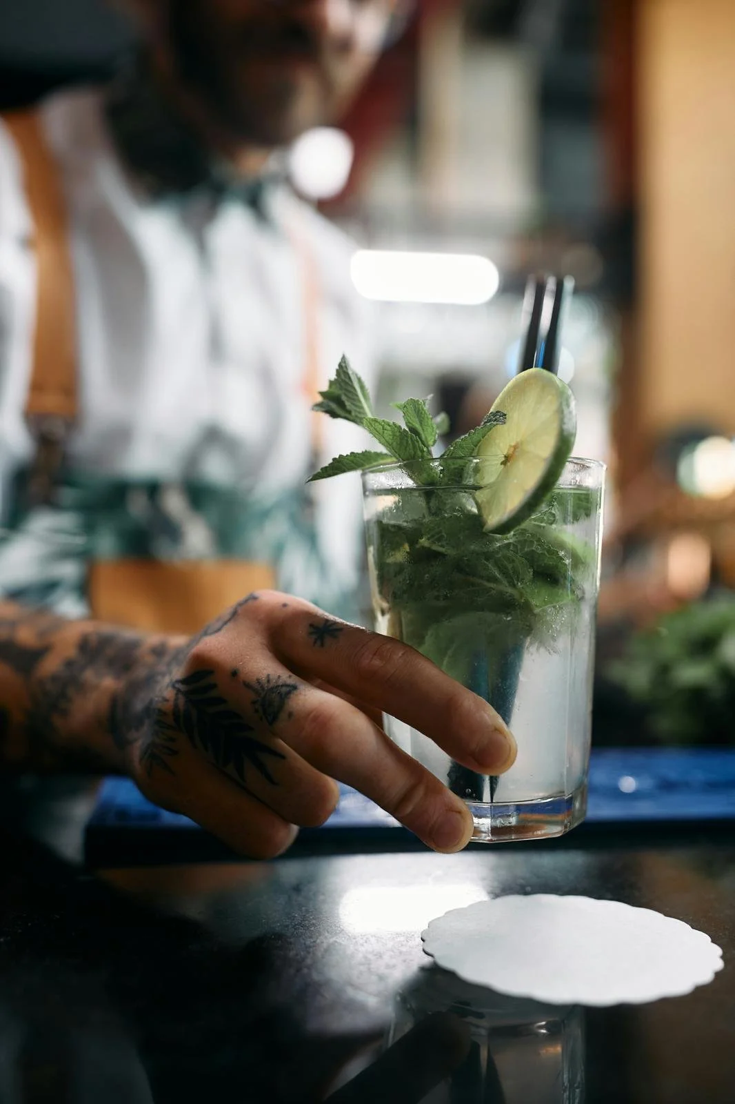 Close-up of a tattooed hand holding a mojito cocktail with lime wedge and mint leaves, on a dark bar counter.