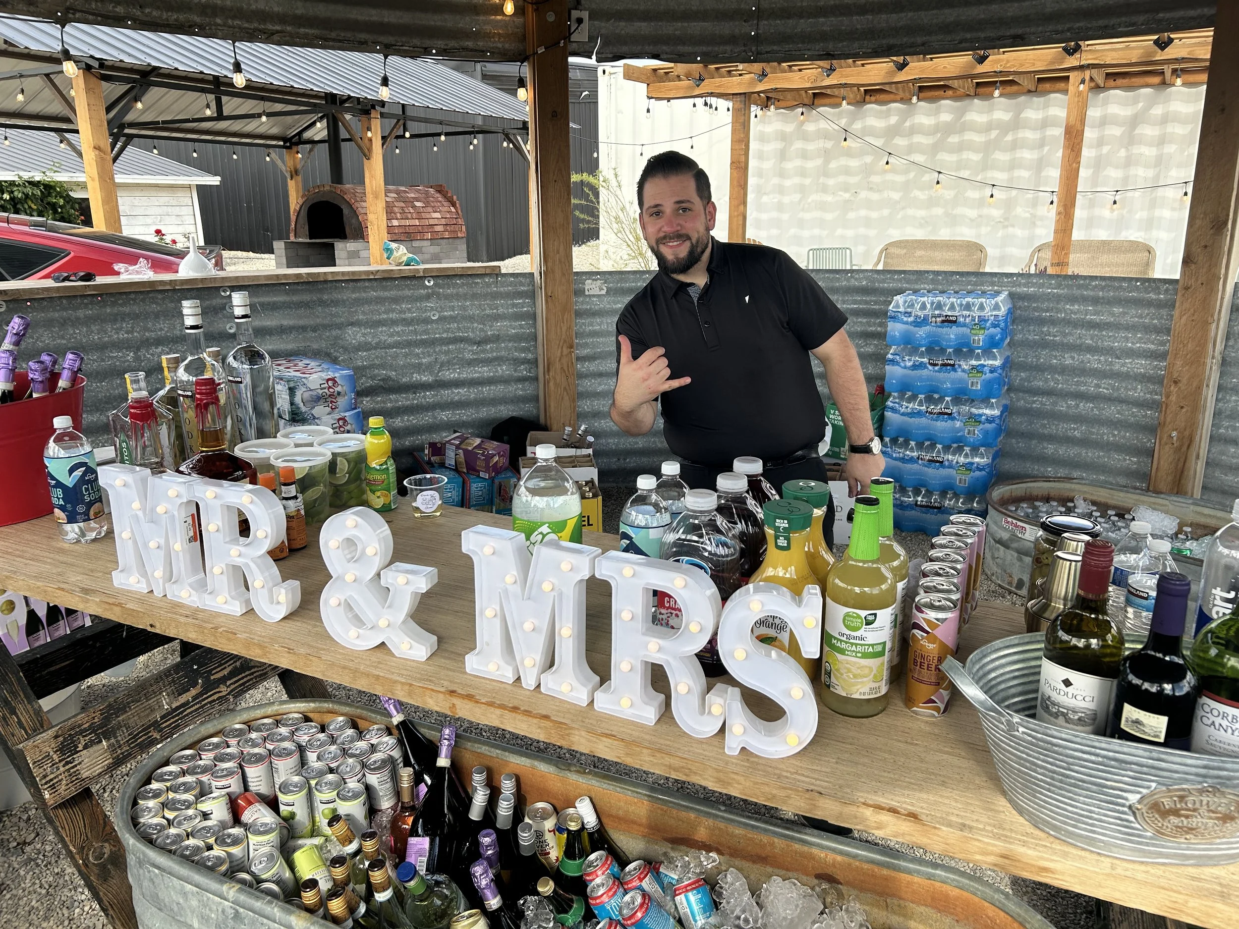 A man with a beard making a shaka sign at a bar setup with various drinks, illuminated decorative letters, and bottled beverages on a wooden table at an outdoor event.
