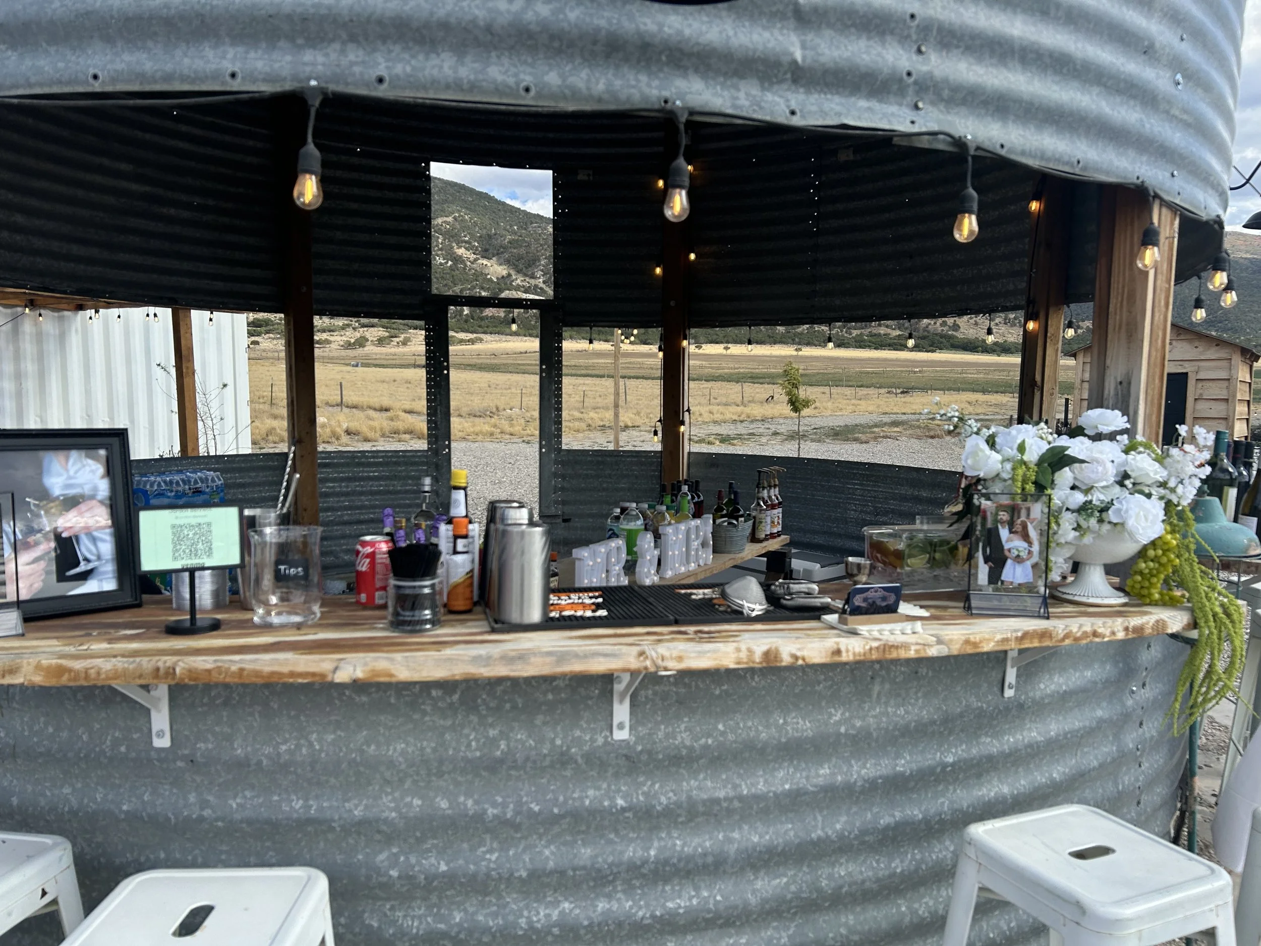 A rustic outdoor bar with a corrugated metal exterior, set up in a rural field with mountains in the background. The bar counter is made of wood and decorated with flowers, framed pictures, and various bottles and bar supplies, with string lights hanging from the roof.