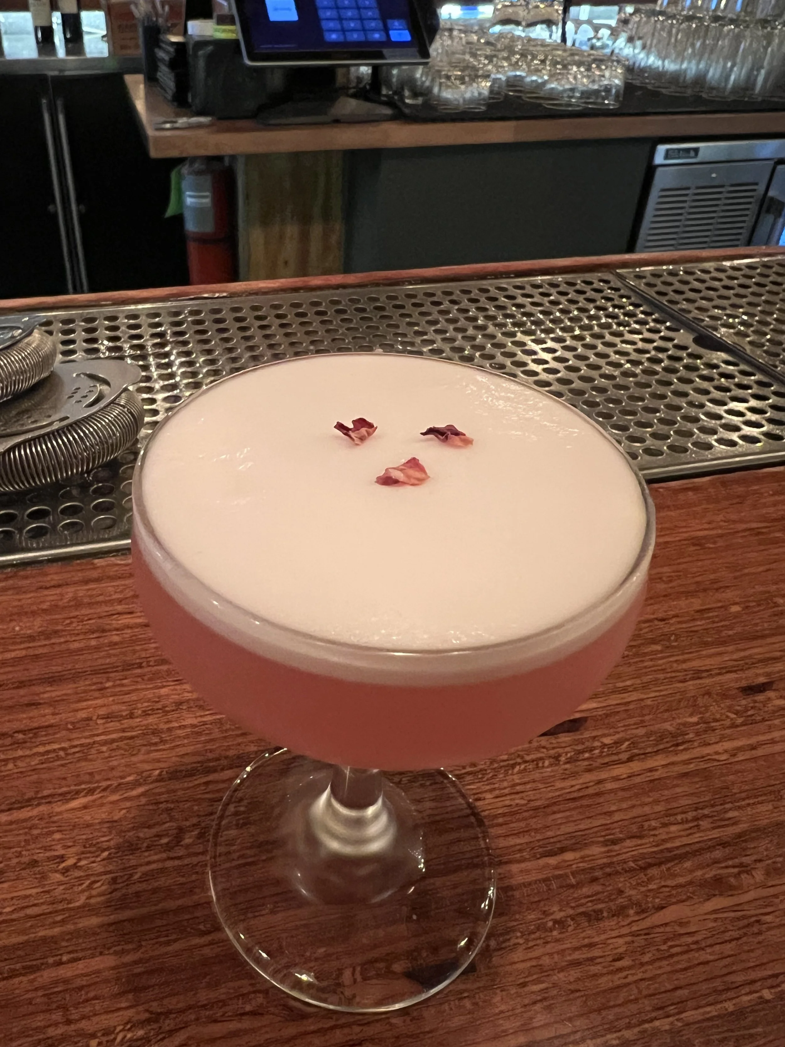 A pink cocktail in a coupe glass with a frothy white top and small flower petals garnish, placed on a wooden bar counter.