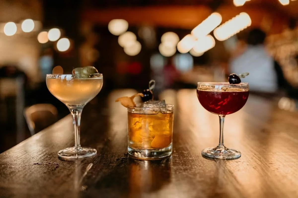 Three cocktails on a wooden bar counter under warm lighting, blurred background with people and hanging lights.