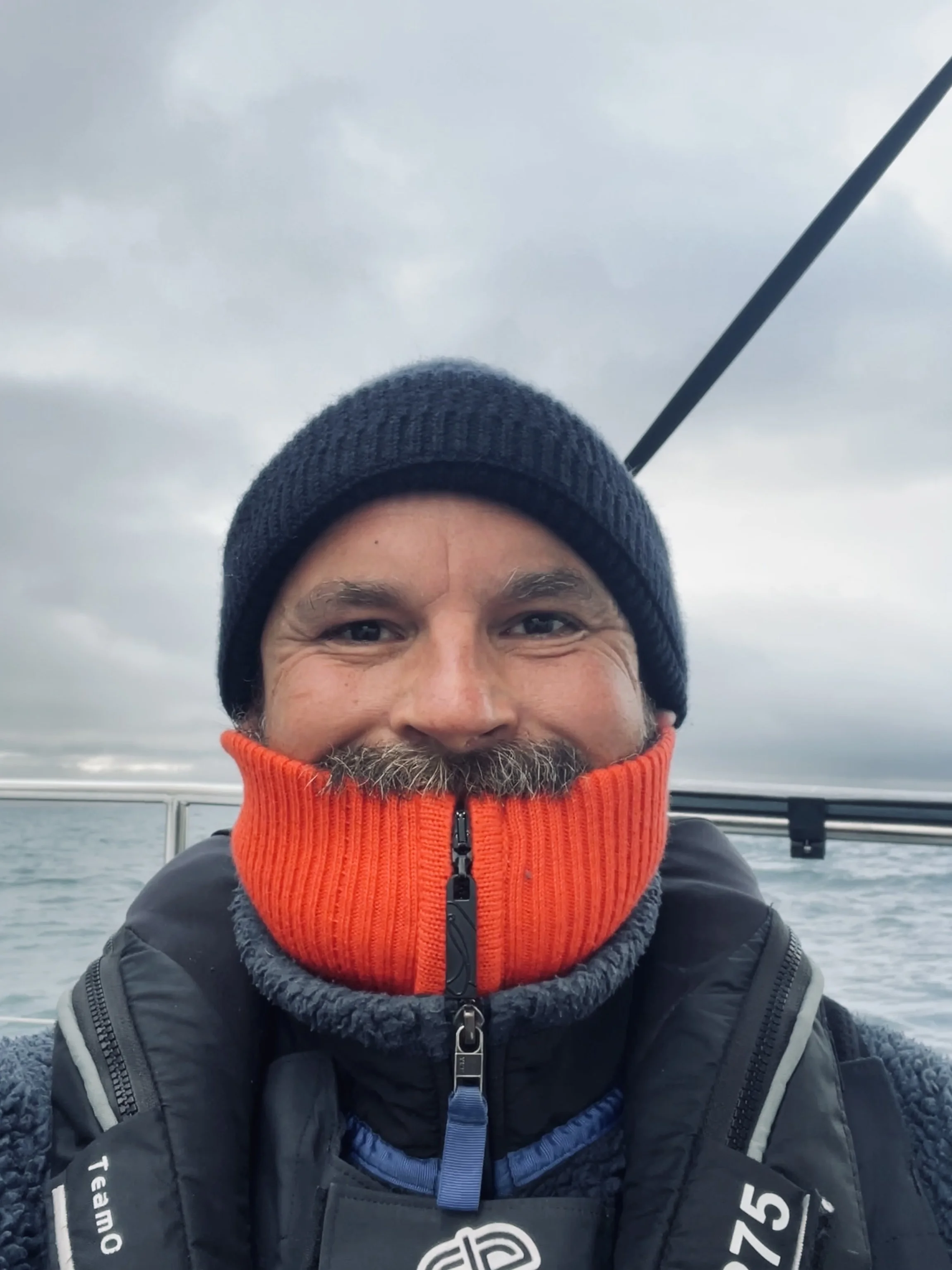 A man with a beard and mustache on a boat, wearing a black beanie and a red and gray fleece zip-up jacket under a black vest. He is smiling with a gray, cloudy sky and water in the background.