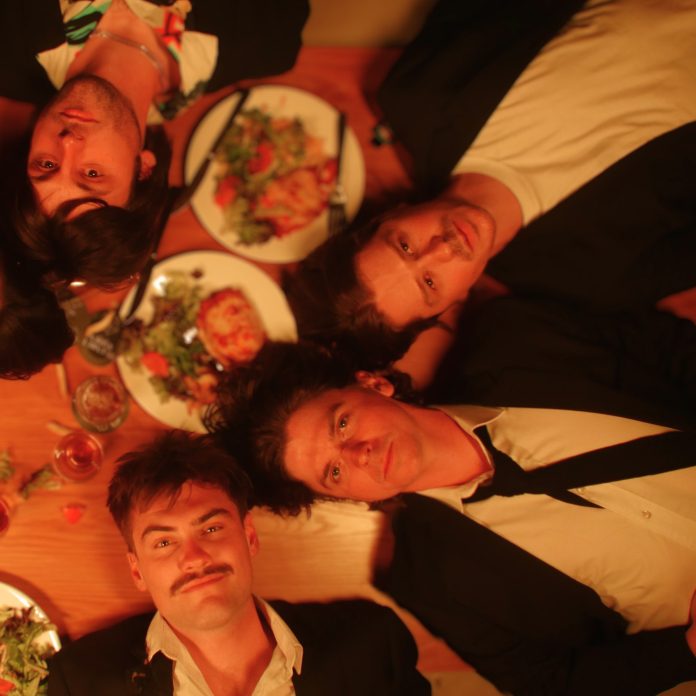 Four people lying on the floor with their heads close together, surrounded by plates of pizza, salad, and drinks at a dinner gathering.