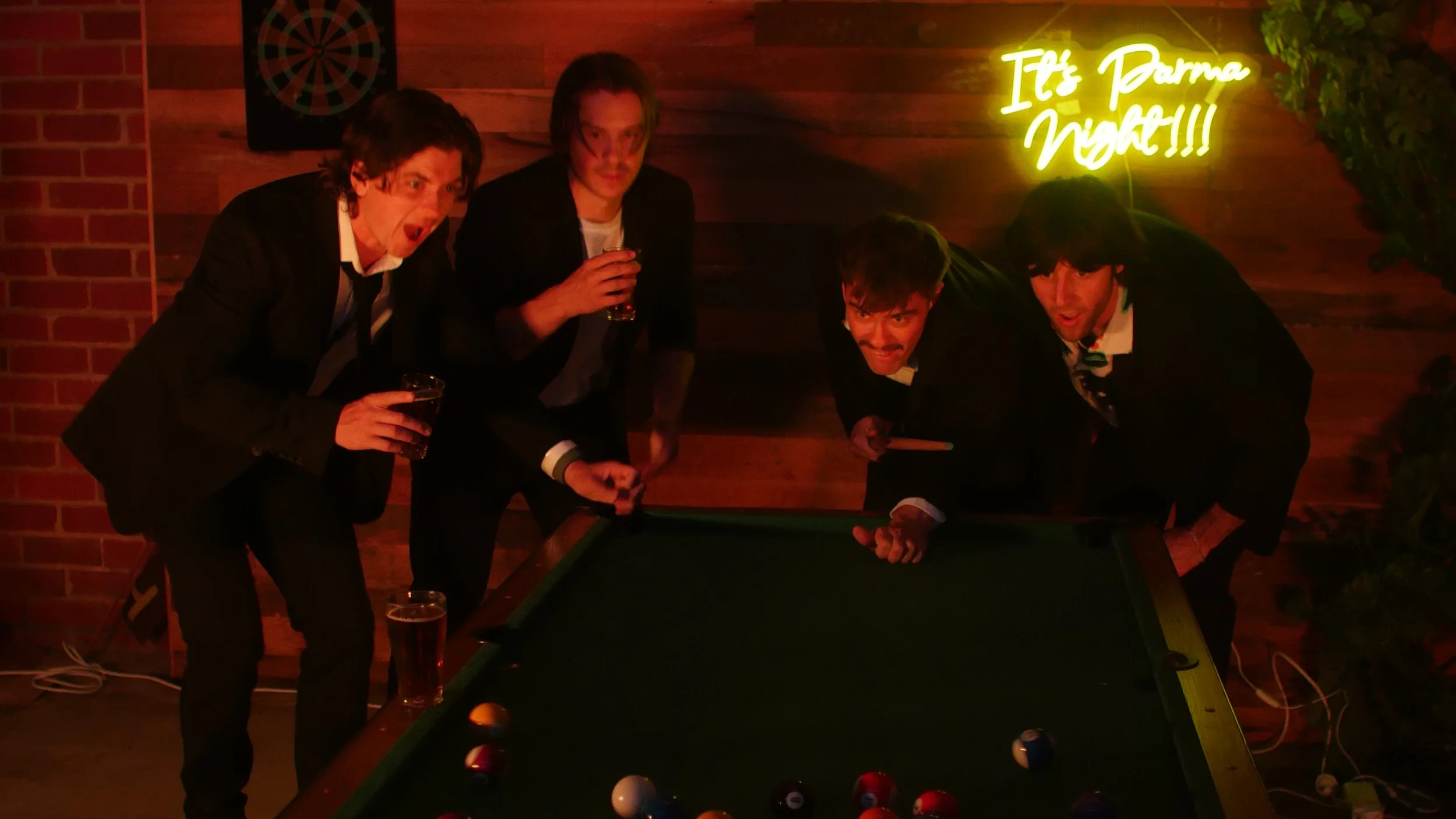 Four men in suits playing pool at a bar with a neon sign that reads 'It's Darma Night!!!' in the background.