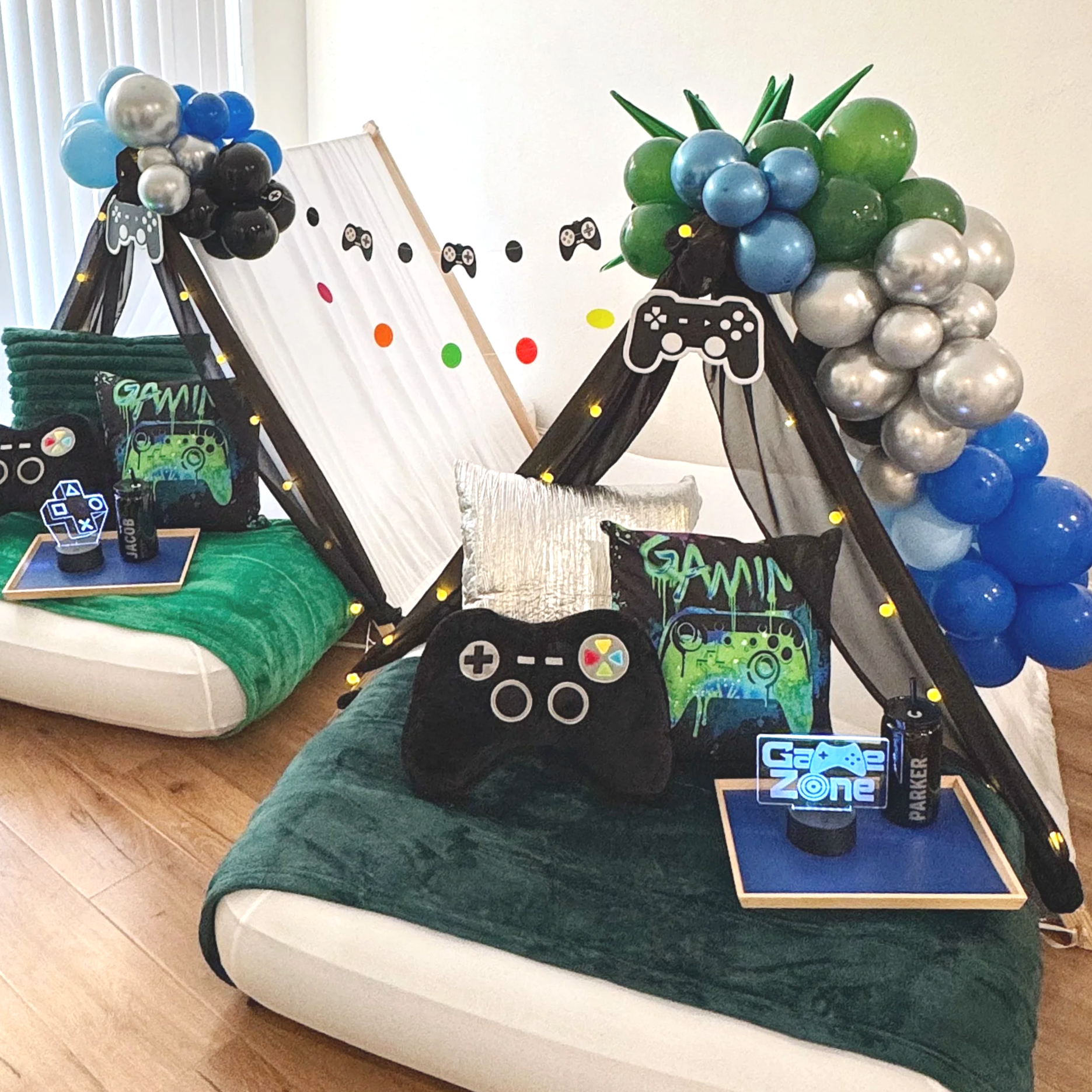 Decorated indoor space with green and black blankets on mattresses, pillows featuring plush gaming controllers for a video game themed party, with balloons arranged decoratively at the top, along with black drapes for a kid's sleepover party.