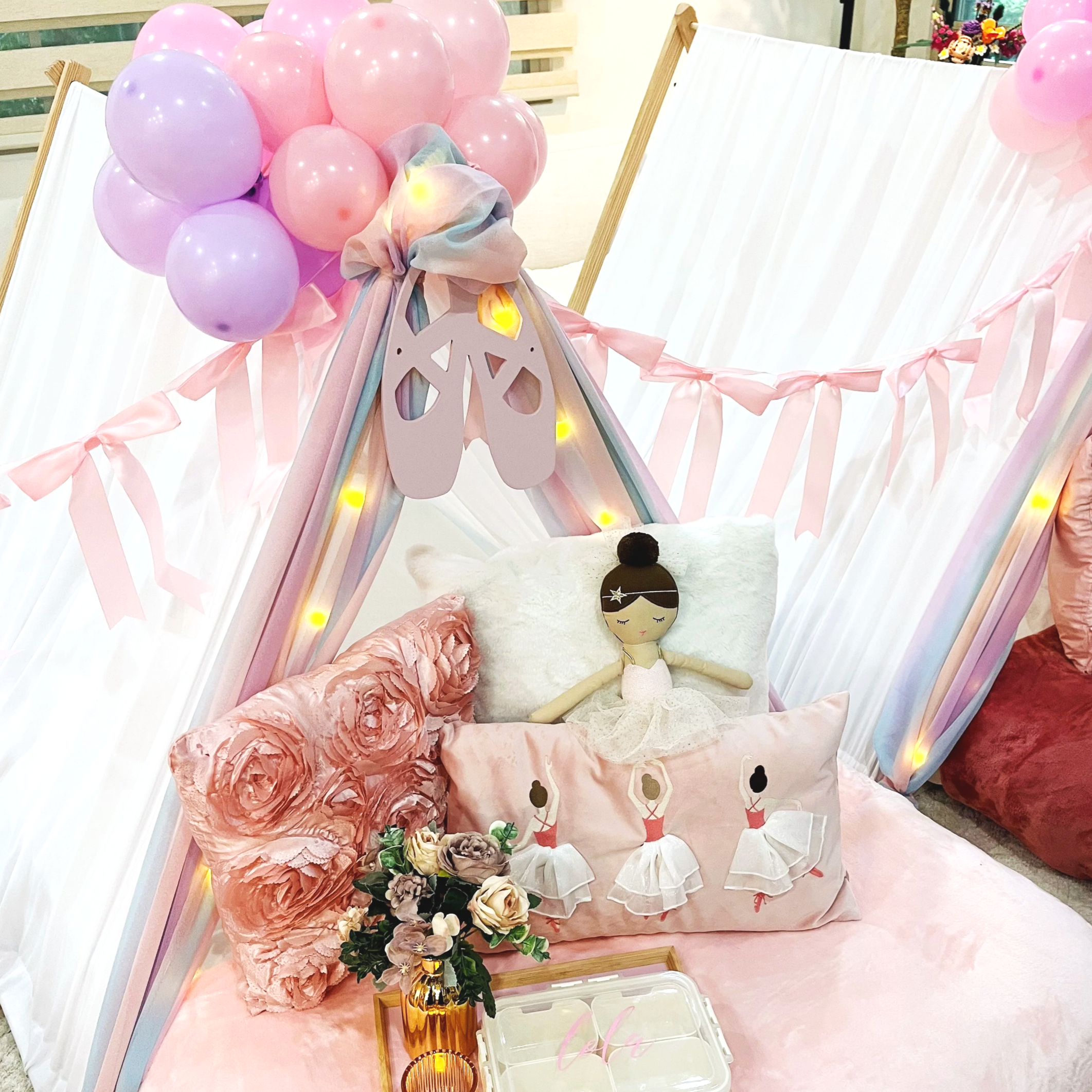 Decorated indoor space with pink blankets on mattresses, plush pillows featuring ballerina characters, and pink and purple balloons arranged decoratively at the top, along with a tray with flowers in a vase and candle for a kid's sleepover party.