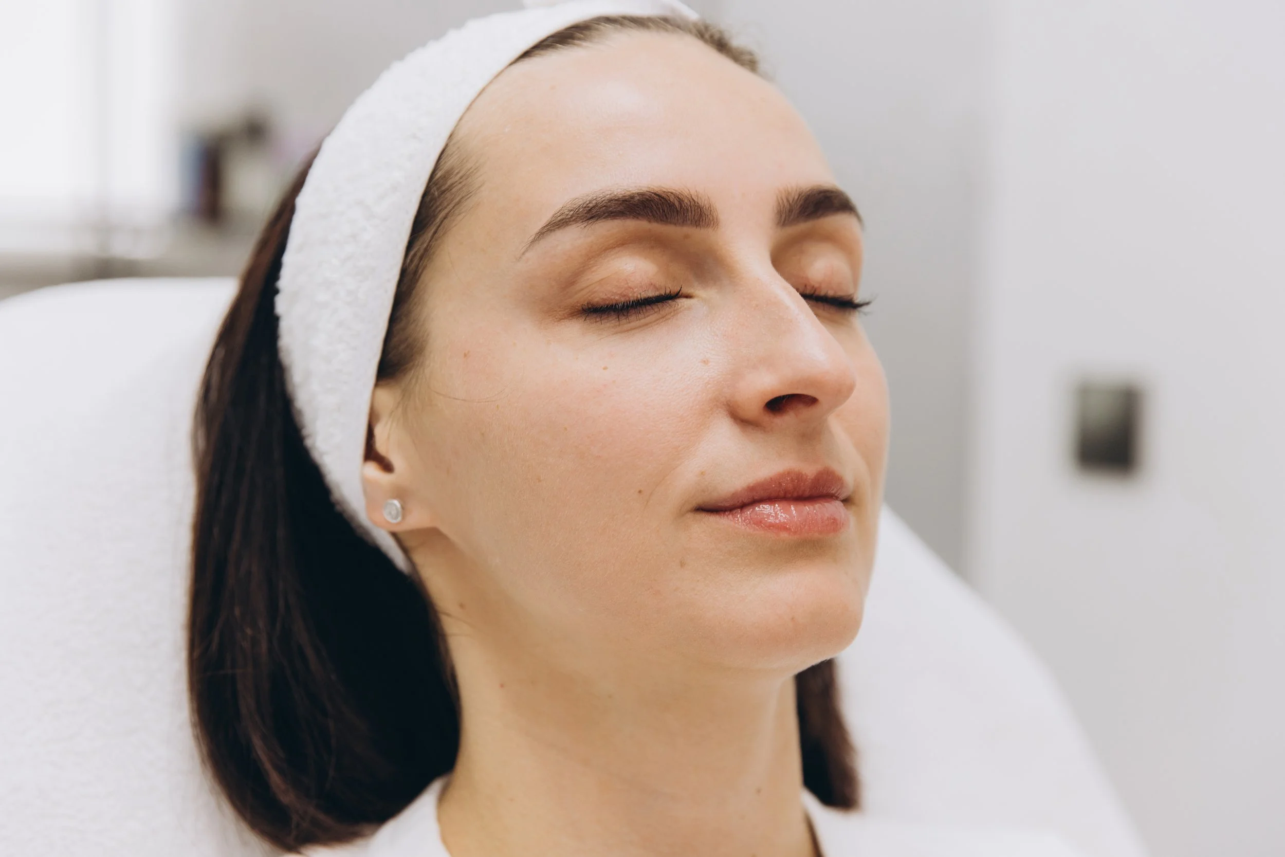 A woman with closed eyes and relaxed facial expression, wearing a white headband and small stud earrings, sitting in a treatment room.