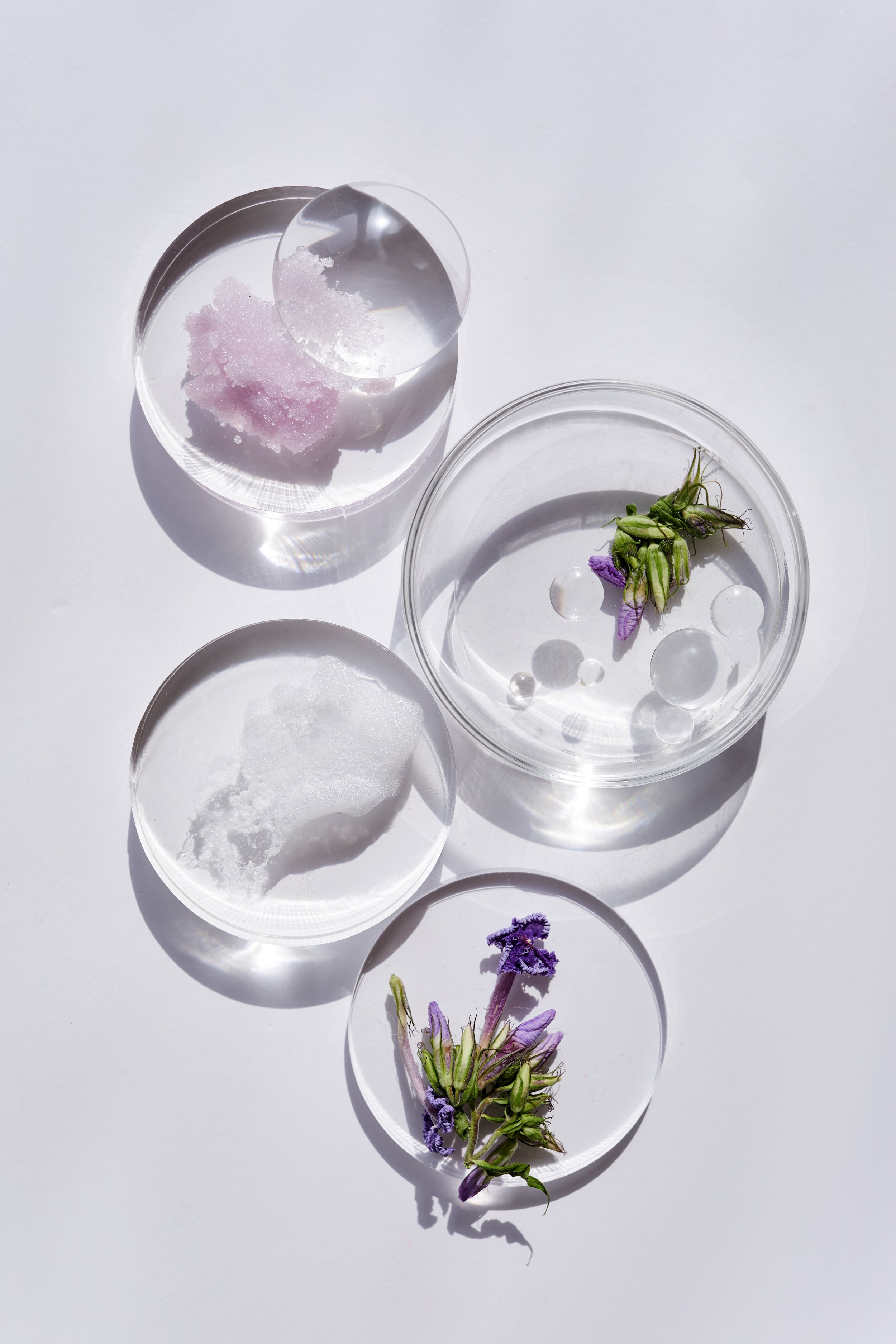 Arrangement of fashion or skincare products in clear glass containers on a white surface, including various transparent and white gels, stones, and small purple and green flowers.