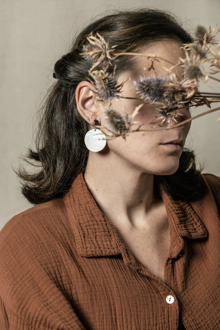Une femme portant une chemise marron, avec des boucles d'oreilles blanches en porcelaine, conçue par l'atelier Moca. Sa tête est inclinée et elle est partiellement voilée par des fleurs séchées.