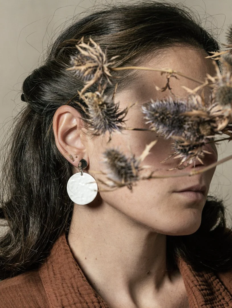 Portrait d'une femme portant un vêtement marron, avec une tige de plante sèche devant son visage. Elle porte des boucles d'oreilles en porcelaine, conçue par l'atelier Moca.