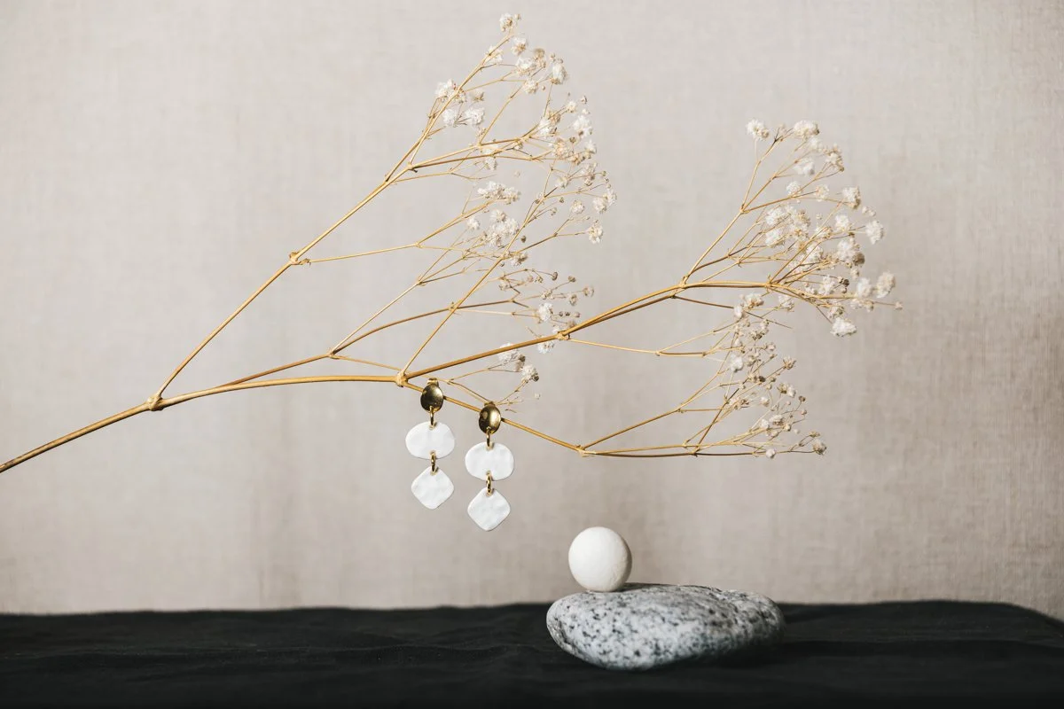 Une paire de boucles d'oreilles blanches en porcelaine, conçue par l'atelier Moca, suspendues à une branche sèche avec de petites fleurs blanches. En dessous, une pierre plate et une boule blanche, sur une surface sombre.
