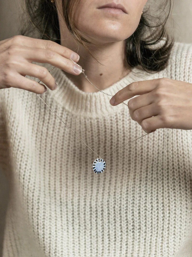 Une femme tenant un collier en argent avec un pendentif rond et bleu ciel, portant un pull en maille beige.