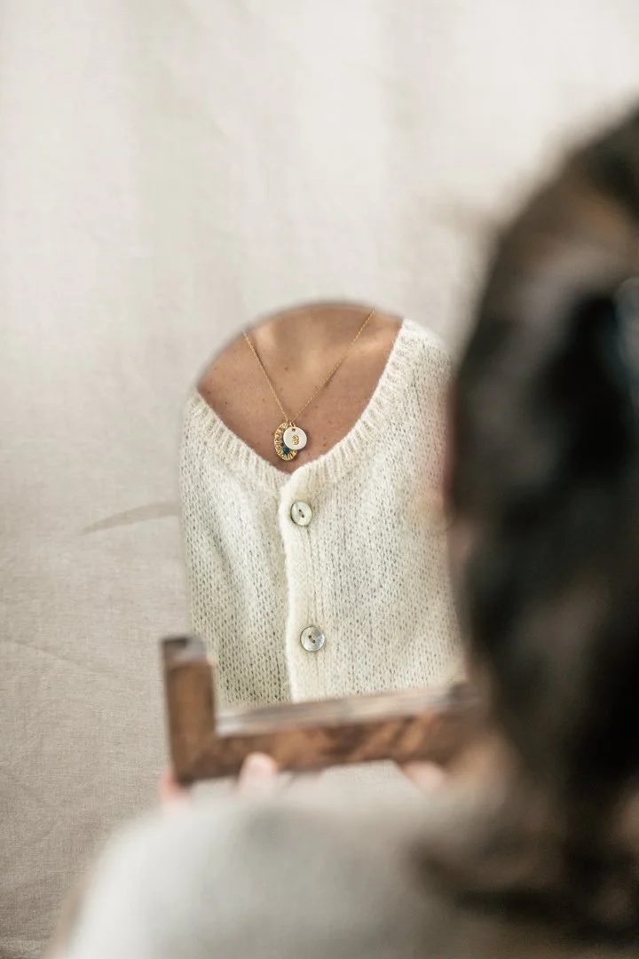 Personne regardant dans un miroir, portant un collier avec un pendentif rond en or et un pull beige à boutons.