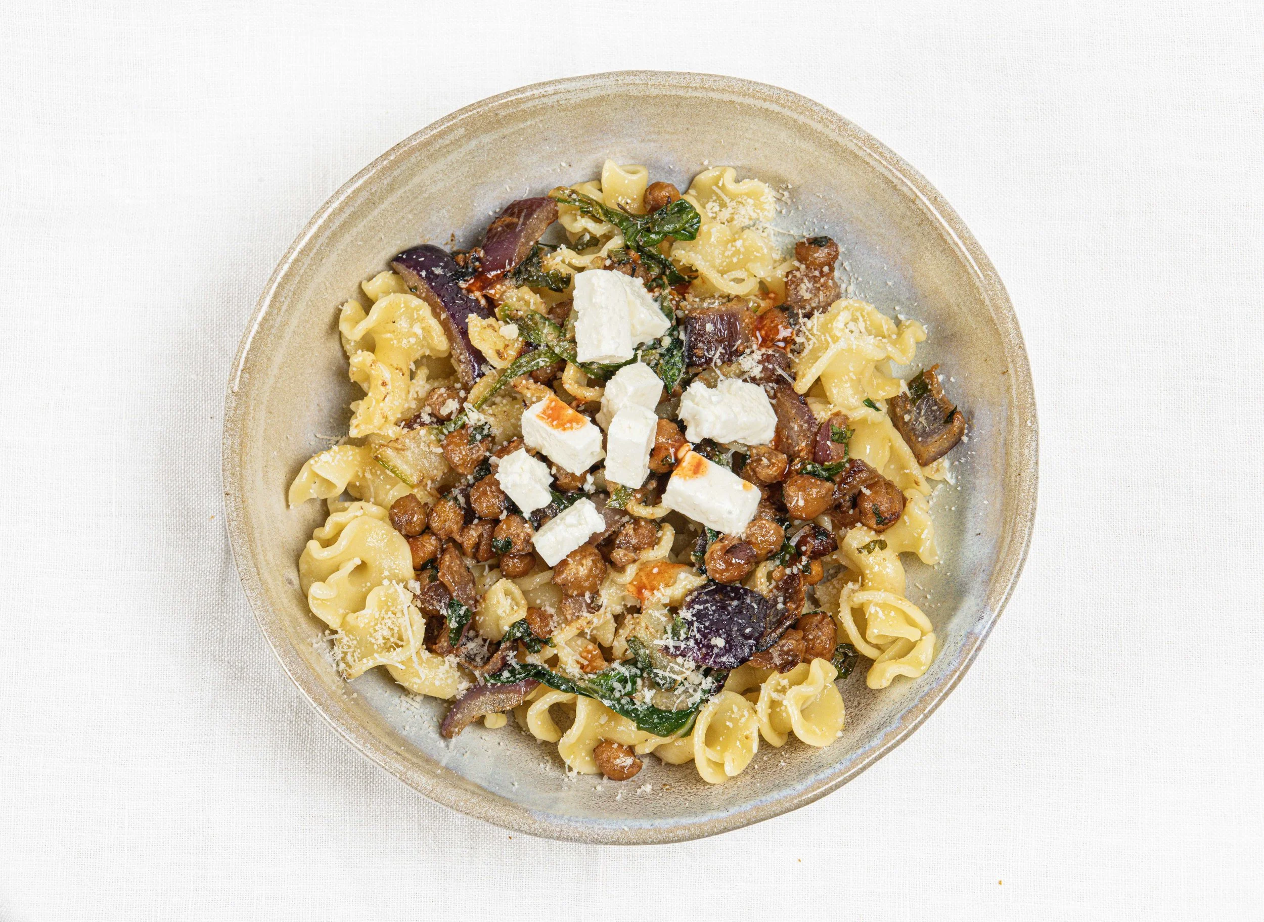 Assiette de pâtes farfalle avec des légumes grillés, des cubes de fromage feta, et des lentilles, servie sur une table blanche. Par le photographe culinaire Yves Daniel. 