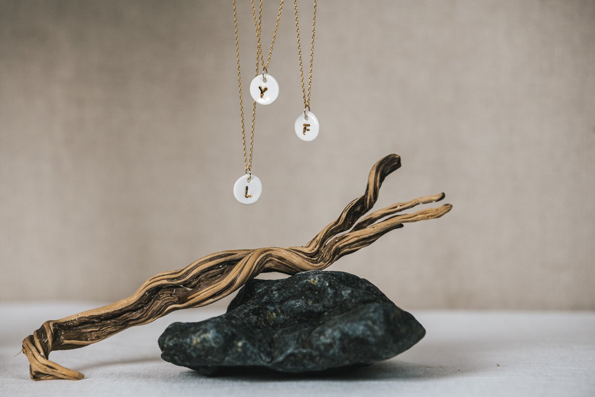 Three necklaces avec des pendentifs en forme de cercles blancs portant les lettres Y, F, L, suspendus au-dessus d'une branche de bois reposant sur une pierre noire.