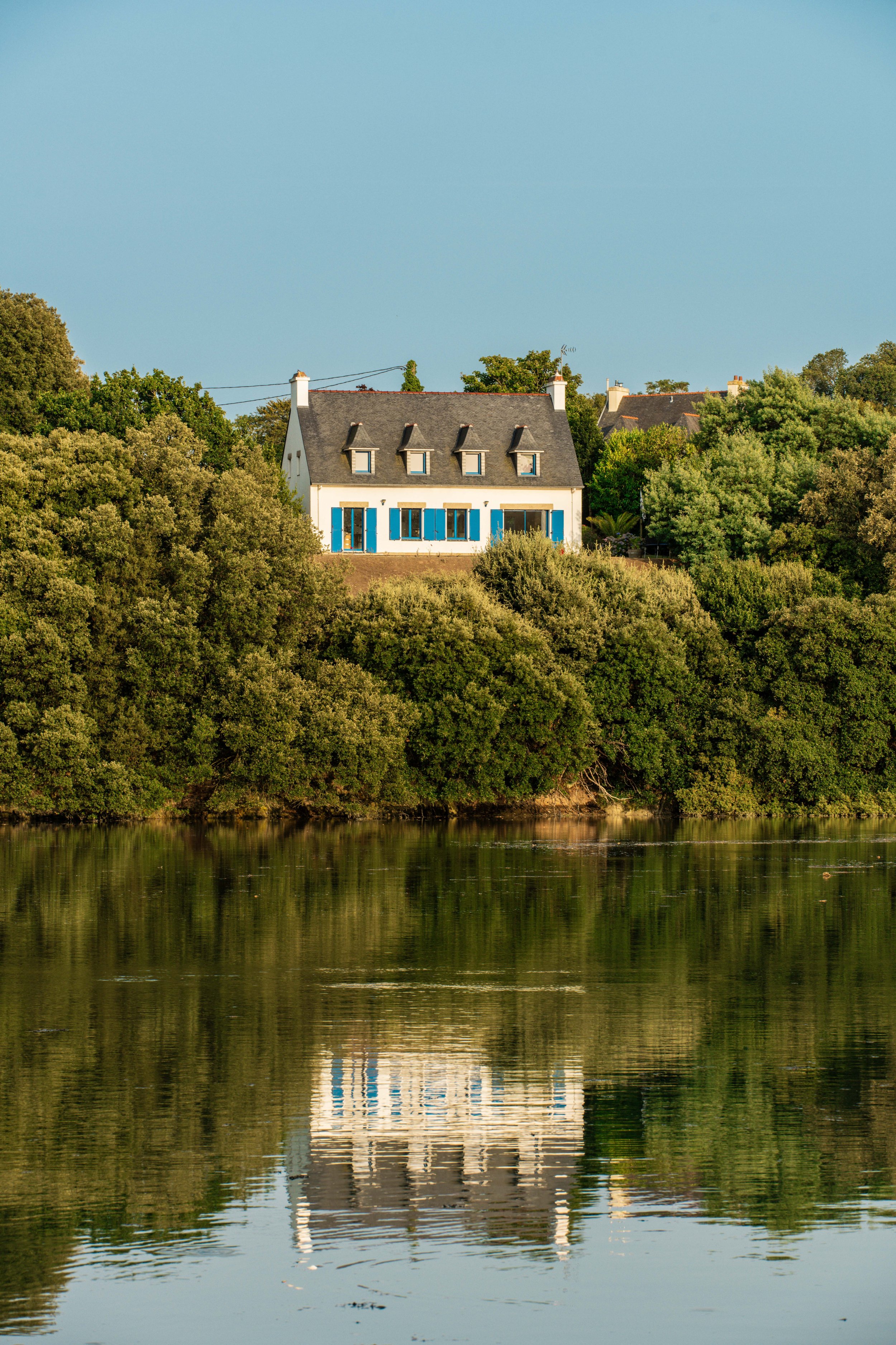 Une maison blanche avec des fenêtres bleues, située sur une colline verdoyante au bord d'une rivière, avec un ciel clair en arrière-plan.