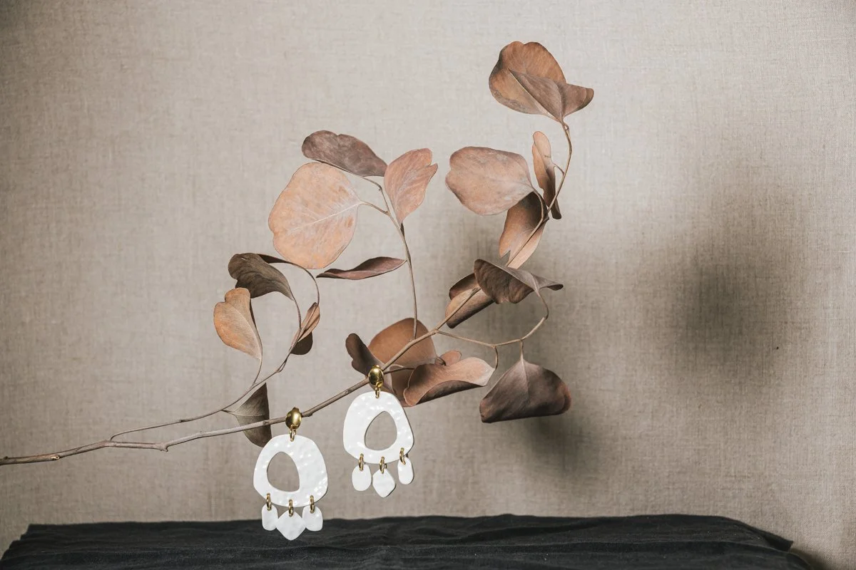 Une paire de boucles d'oreilles blanches en porcelaine, conçue par l'atelier Moca, suspendues à une branche avec des feuilles brunes sur un fond neutre.