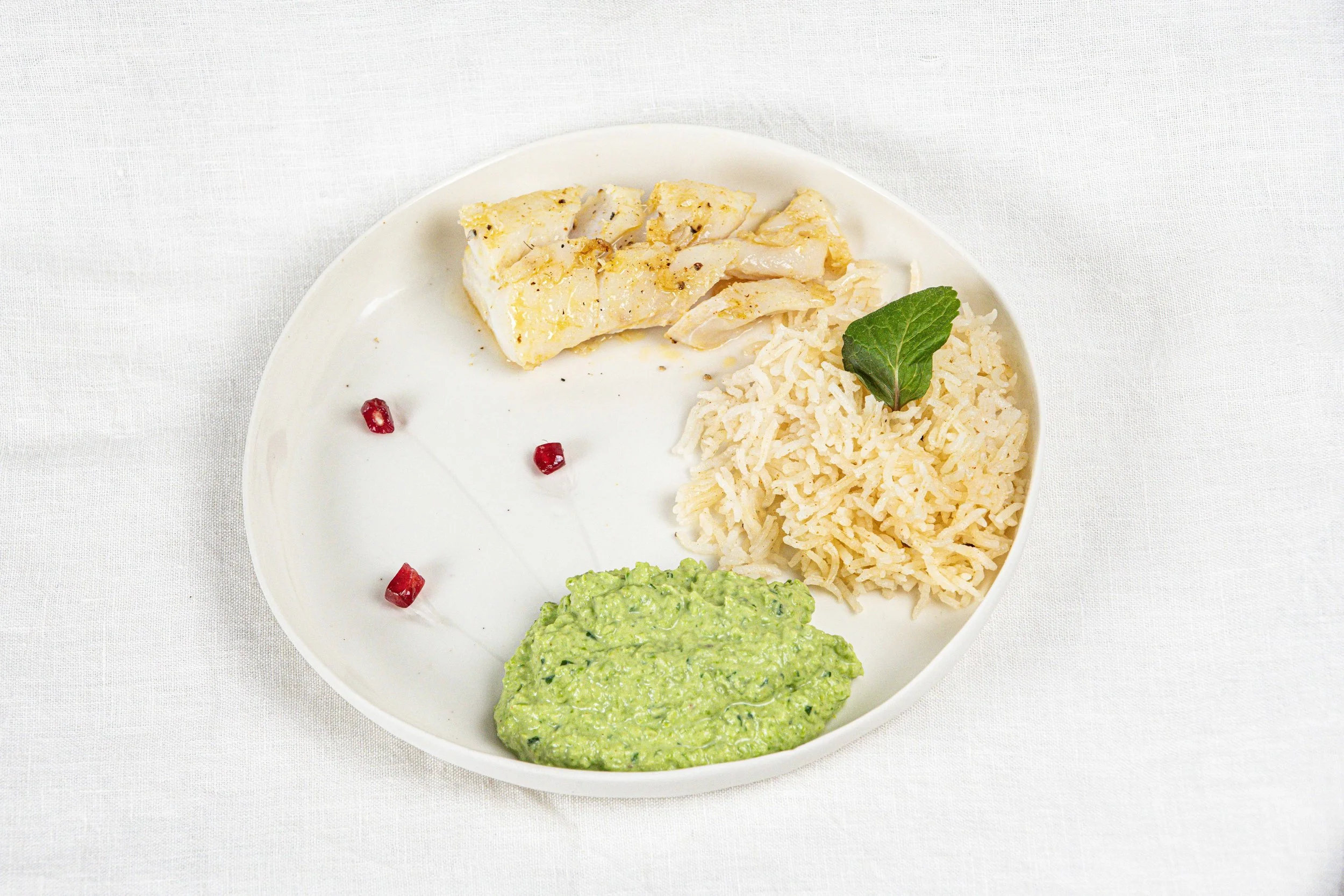 Assiette avec du poisson blanc, du riz blanc, une sauce verte, et quelques graines de grenade, décorée d'une feuille de menthe, sur fond blanc. Par le photographe culinaire Yves Daniel. 