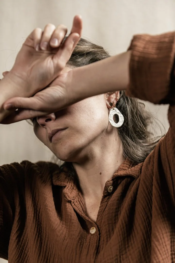 Une femme avec des boucles d'oreilles en forme d'anneau, conçue par l'atelier Moca, portant une chemise brune en velours côtelé, couvre son visage avec sa main dans une pose artistique.