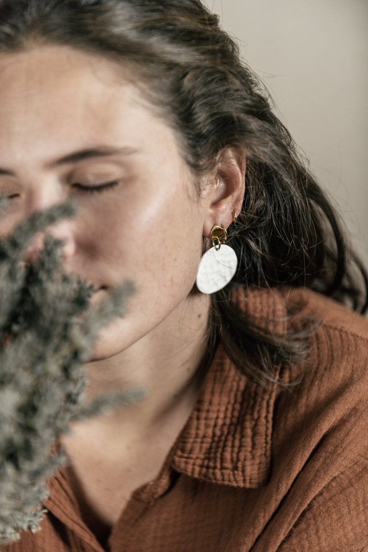 Jeune femme aux yeux fermés, habillée d'une veste marron, près d'une plante floue. Elle porte des boucles d'oreilles en porcelaine, conçue par l'atelier Moca.