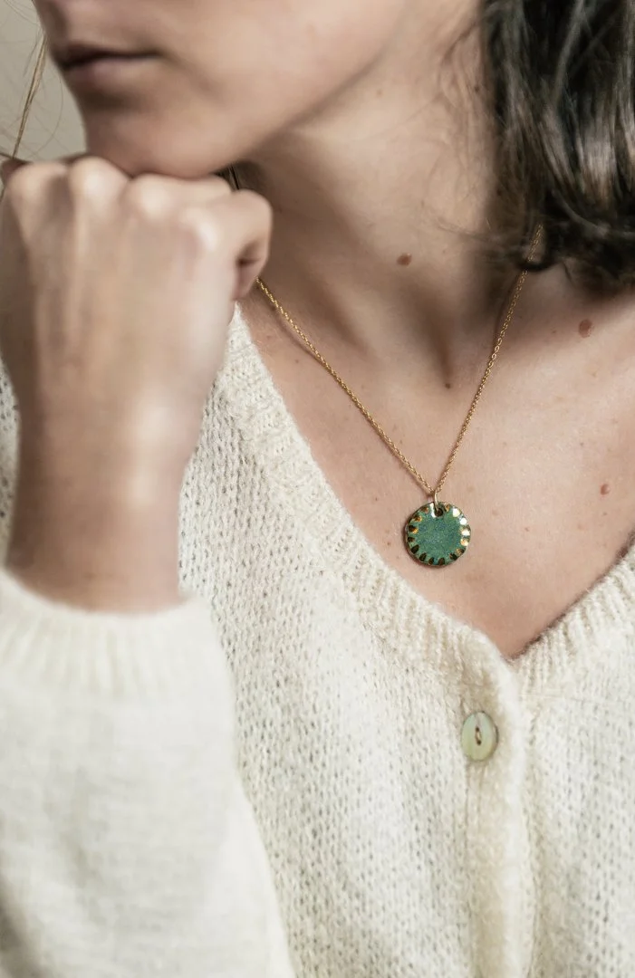 Une femme portant un pull beige et un collier avec un pendentif circulaire vert avec des détails dorés, conçue par l'atelier Moca.