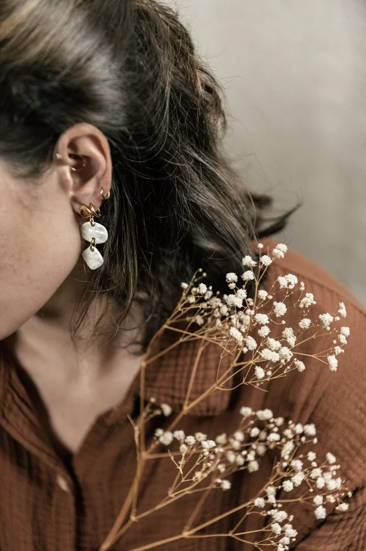 Une femme porte des boucles d'oreilles en porcelaine, conçu par l'atelier Moca, et tient un bouquet de petites fleurs blanches.