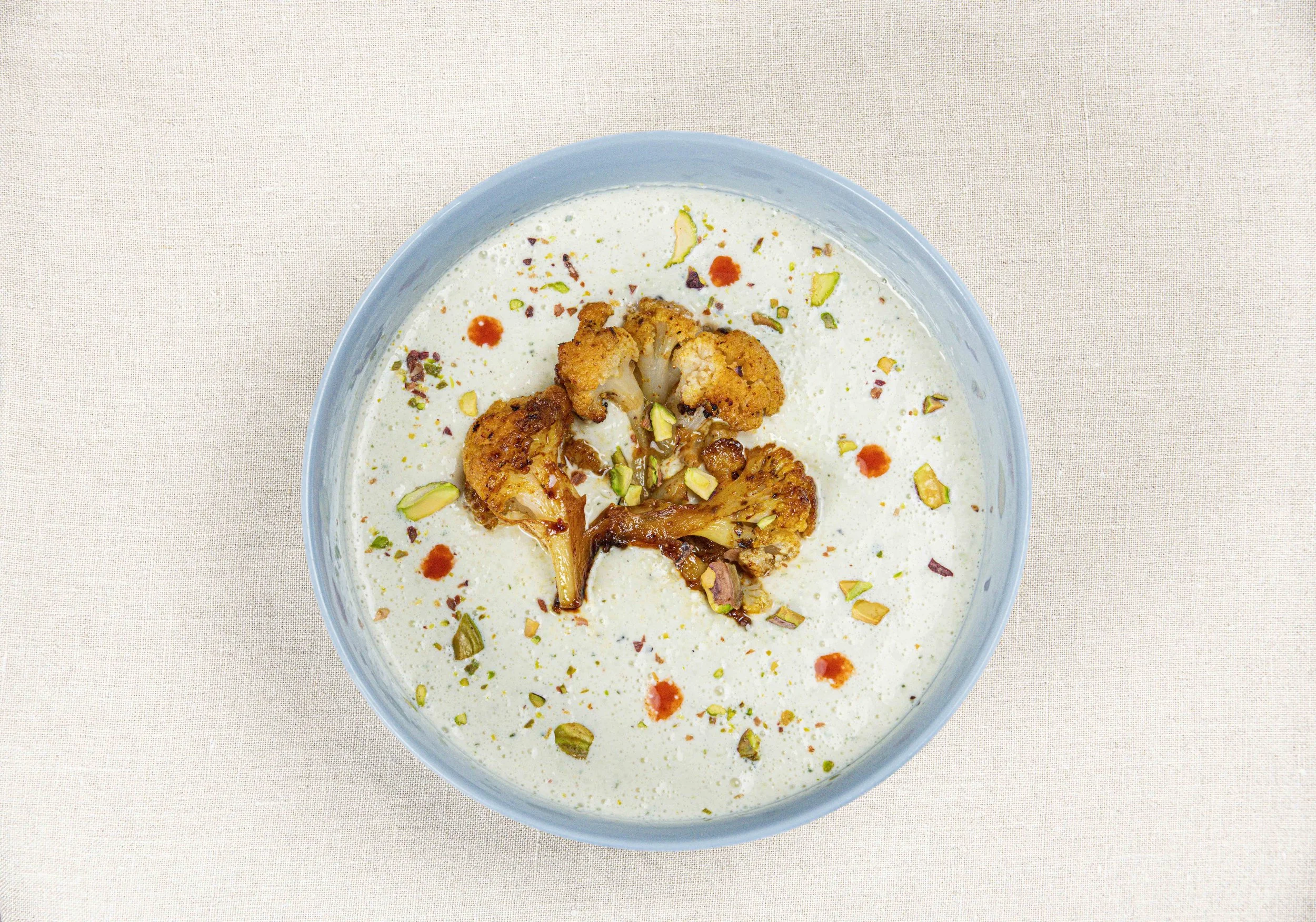 Soupe de chou-fleur garnie de florets de chou-fleur frits et de pistaches hachées, servie dans un bol bleu sur une nappe en lin beige. Par le photographe culinaire Yves Daniel. 