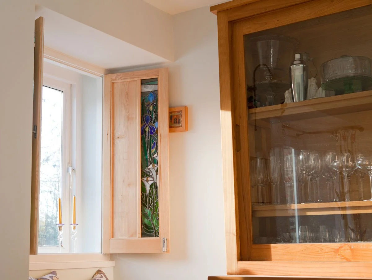 Interior view of a room with a window, a wooden cabinet with stained glass, a small framed picture on the wall, and a glass-fronted cabinet with glassware inside.