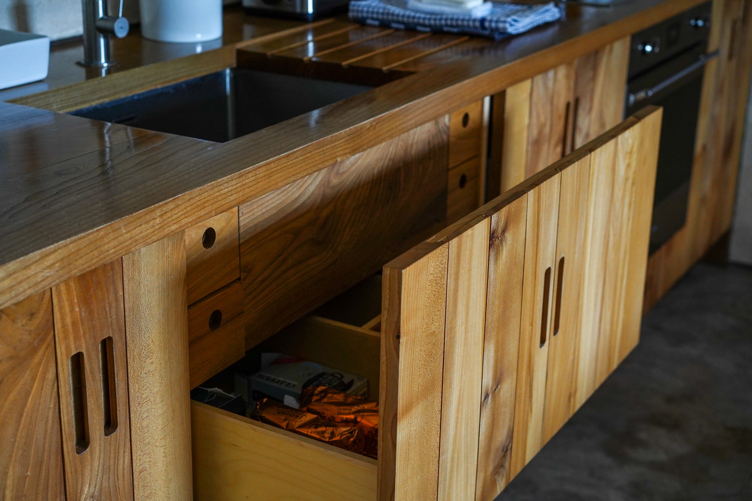 A wooden kitchen cabinet with one door open, revealing an interior lower drawer with some items inside. The countertop has a dark sink, a white item, and a dishcloth.