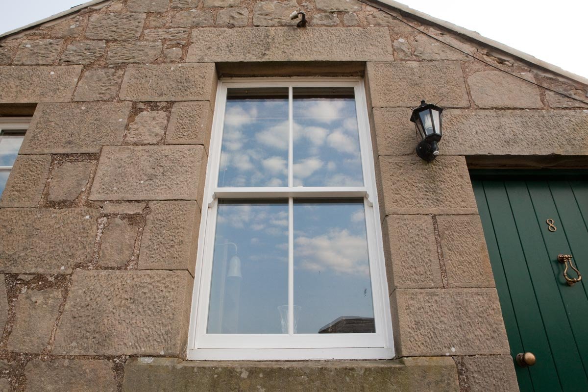 A stone house wall with a window reflecting clouds and sky, a black outdoor lantern next to the window, and a green door with the number 8 and a brass knocker.