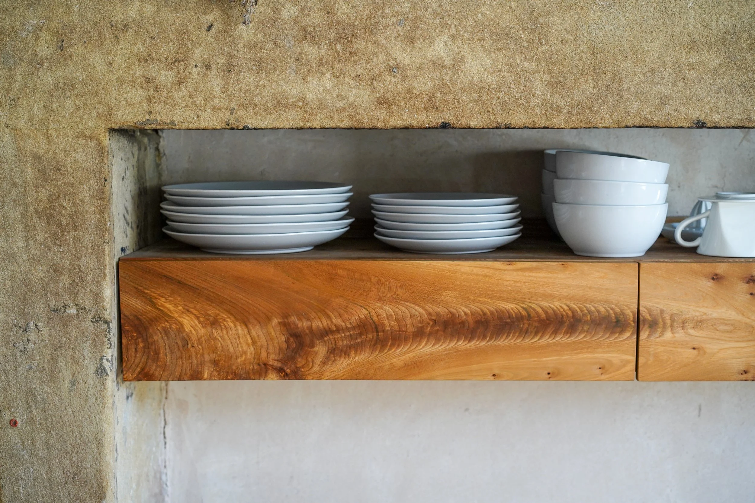 White dishes and bowls arranged on a wooden shelf within a rustic wall opening.