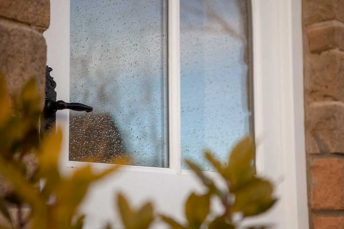 Rain on a window with a black faucet and brick wall, with blurred yellow leaves in the foreground.