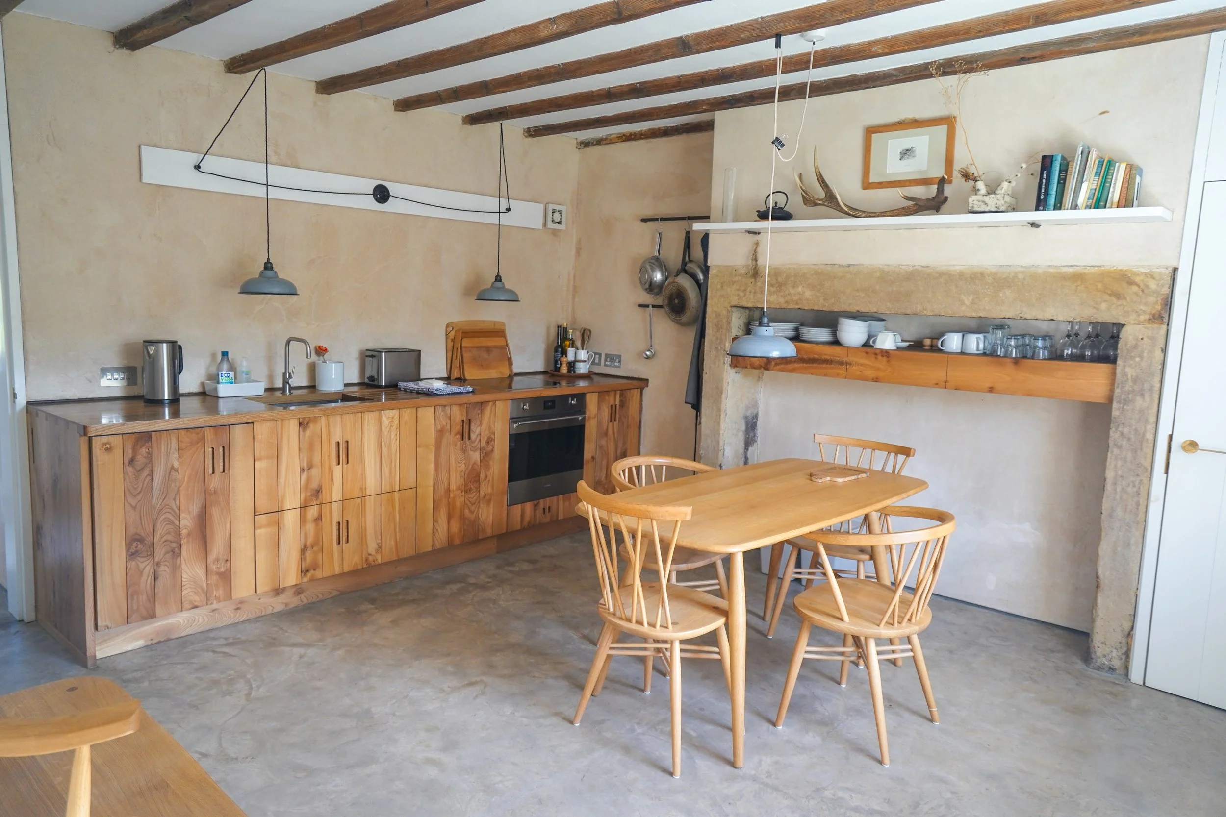 Rustic kitchen with wooden cabinets, a dining table with four chairs, hanging pendant lights, and vintage hanging pans on the wall.