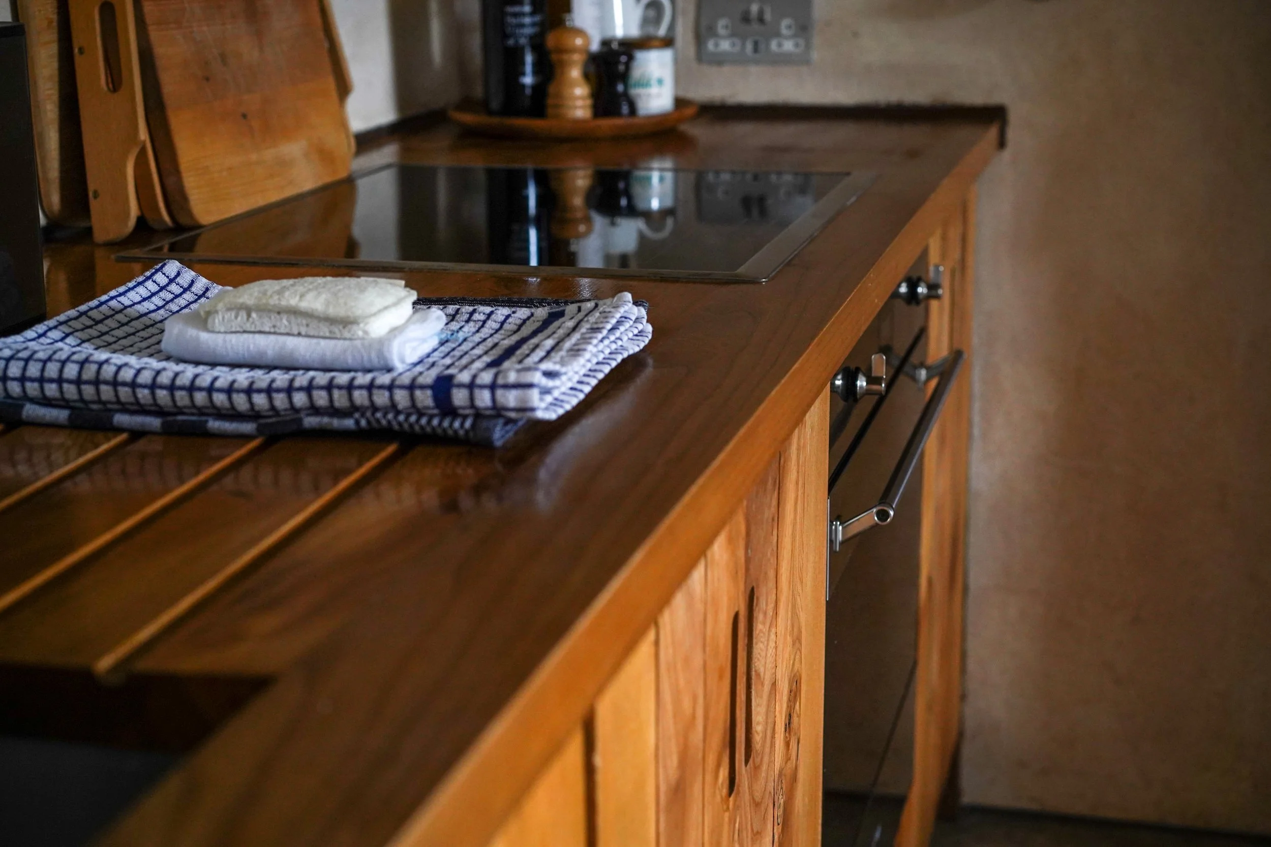 A wooden kitchen countertop with a folded dish towel and a bar of soap on top, near a stovetop and salt and pepper shakers in the background.